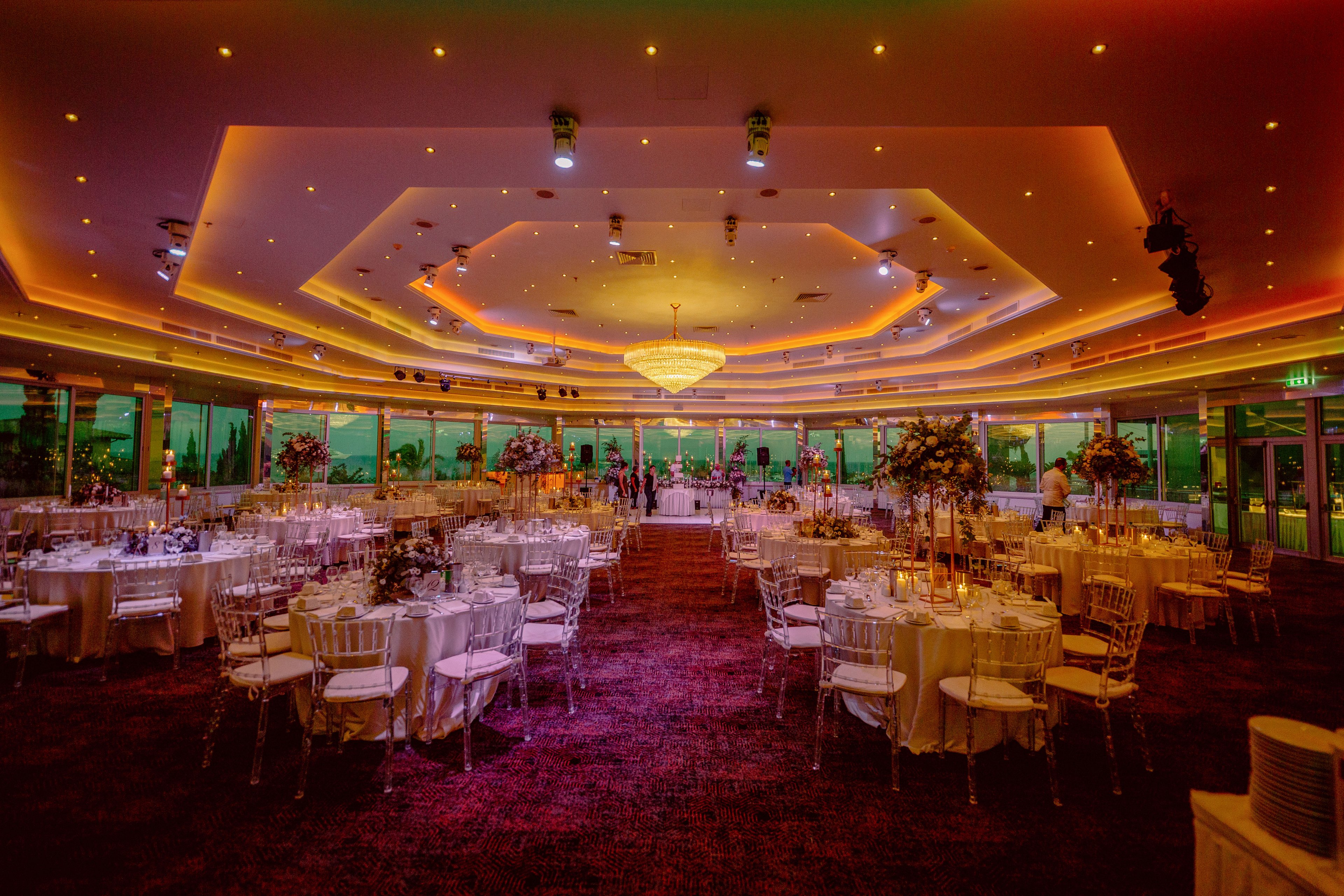 A large, luxurious banquet hall is fully set for an evening reception with round tables and white chairs arranged across the room. Each table is decorated with glassware, candles, and tall floral centerpieces, creating a sophisticated atmosphere. Warm orange and yellow ceiling lights, along with a grand central chandelier, bathe the space in a golden glow. Floor-to-ceiling windows line the back wall, revealing a green-tinted view outside and adding depth to the scene.