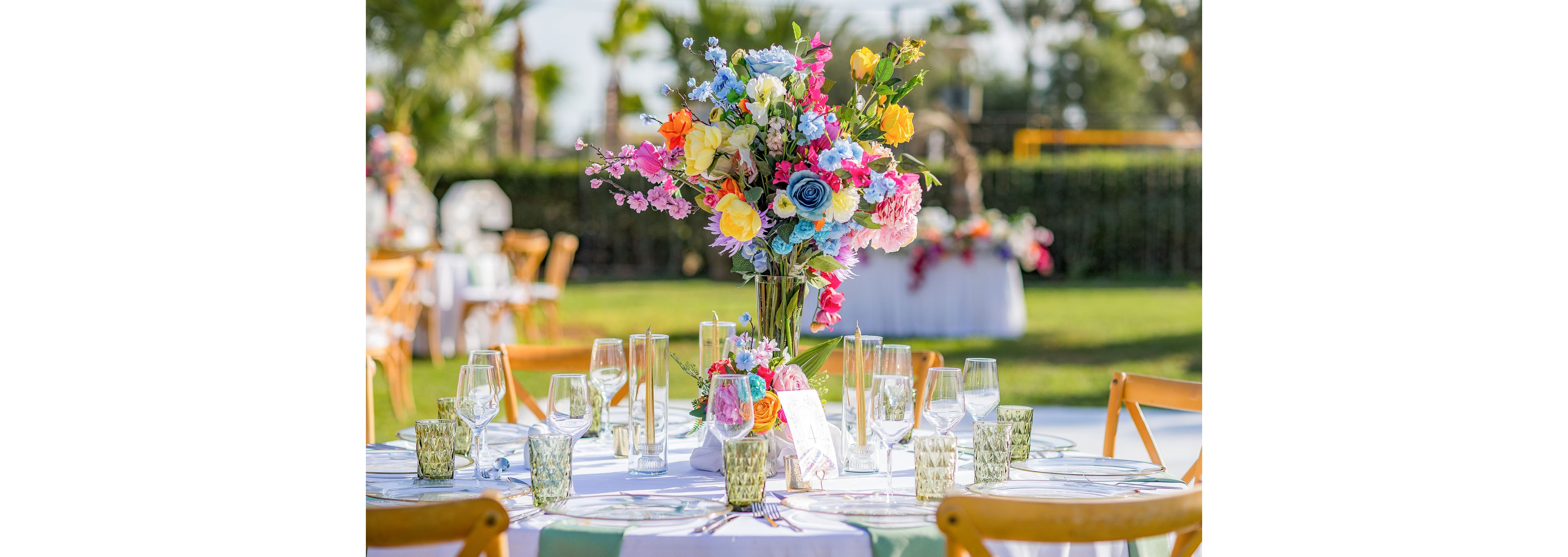 A round table is set outdoors with a tall, colorful floral centerpiece featuring roses and mixed blossoms. Clear glassware, green textured tumblers, and neatly arranged place settings surround the arrangement. Wooden chairs and additional decorated tables are visible in the background, suggesting a festive gathering or wedding reception. The scene is bright and sunny, with greenery and soft-focus guests adding to the celebratory atmosphere.