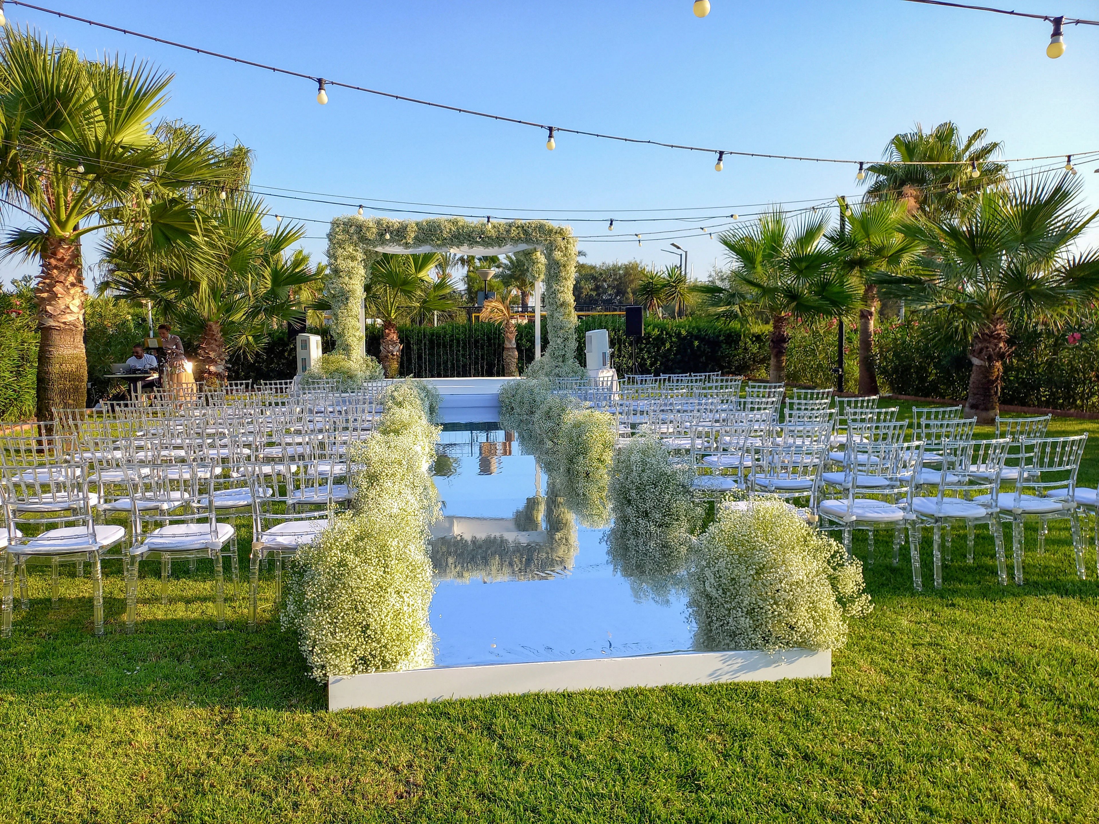 An outdoor wedding ceremony setup features a reflective mirrored aisle lined with lush white baby's breath arrangements. Transparent chairs are arranged in neat rows on either side of the aisle, creating a formal seating area on the green lawn. At the end of the aisle stands a large floral arch covered in white flowers and greenery, framed by palm trees. String lights are suspended overhead, suggesting an event that may extend into the evening under a clear blue sky.