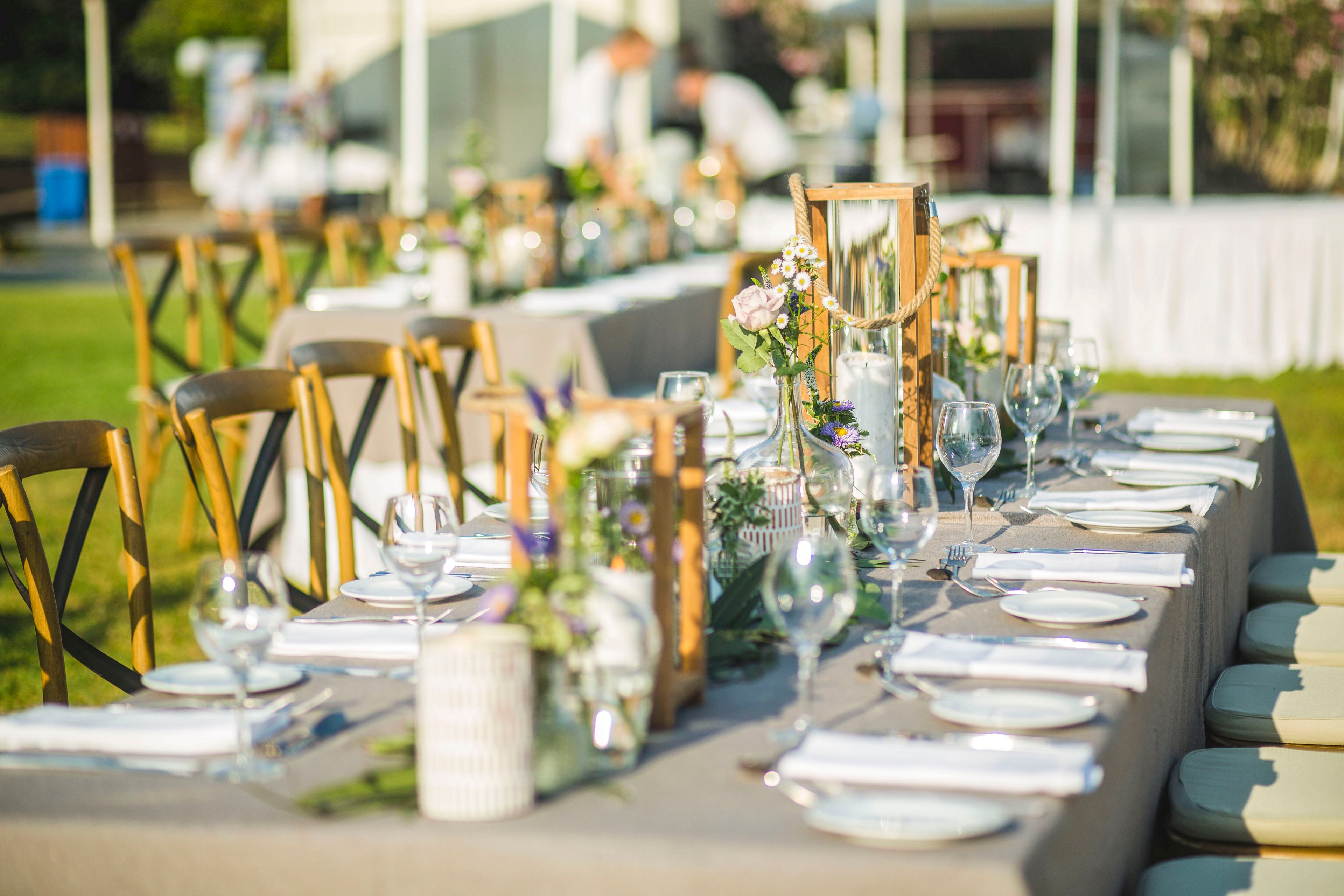 A long banquet table is beautifully arranged for an outdoor event, likely a wedding reception. Wooden chairs line both sides of the table, which is covered with a neutral tablecloth and neatly folded white napkins. Glassware, plates, and cutlery are carefully placed, while floral arrangements and lantern centerpieces add a rustic, romantic touch. The scene is set on a sunny day with a soft, warm color palette and a blurred background of a tented area.