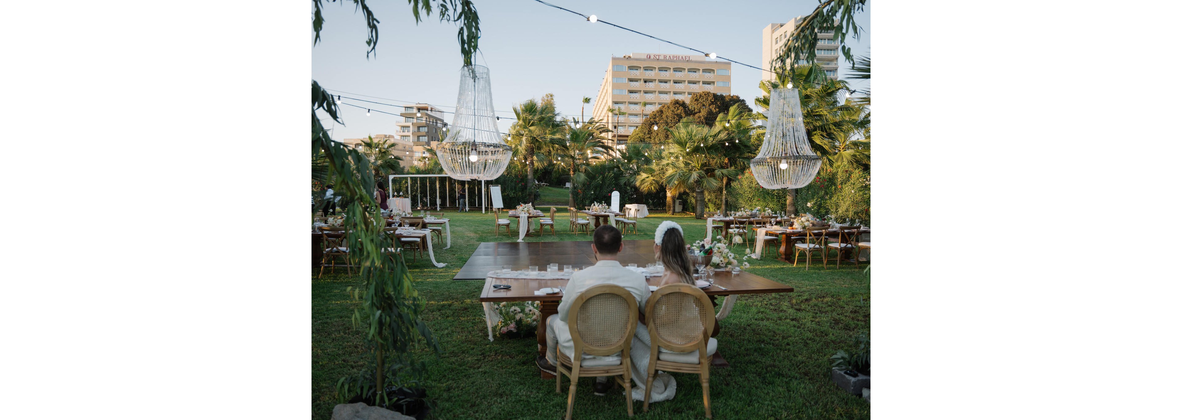A newlywed couple sits together at a long wooden table overlooking an outdoor wedding reception. The setting features a central dance floor surrounded by banquet tables on a lush green lawn with palm trees. Large hanging chandeliers and string lights add an elegant touch to the open-air venue. Modern buildings rise in the background, contrasting with the intimate garden atmosphere.