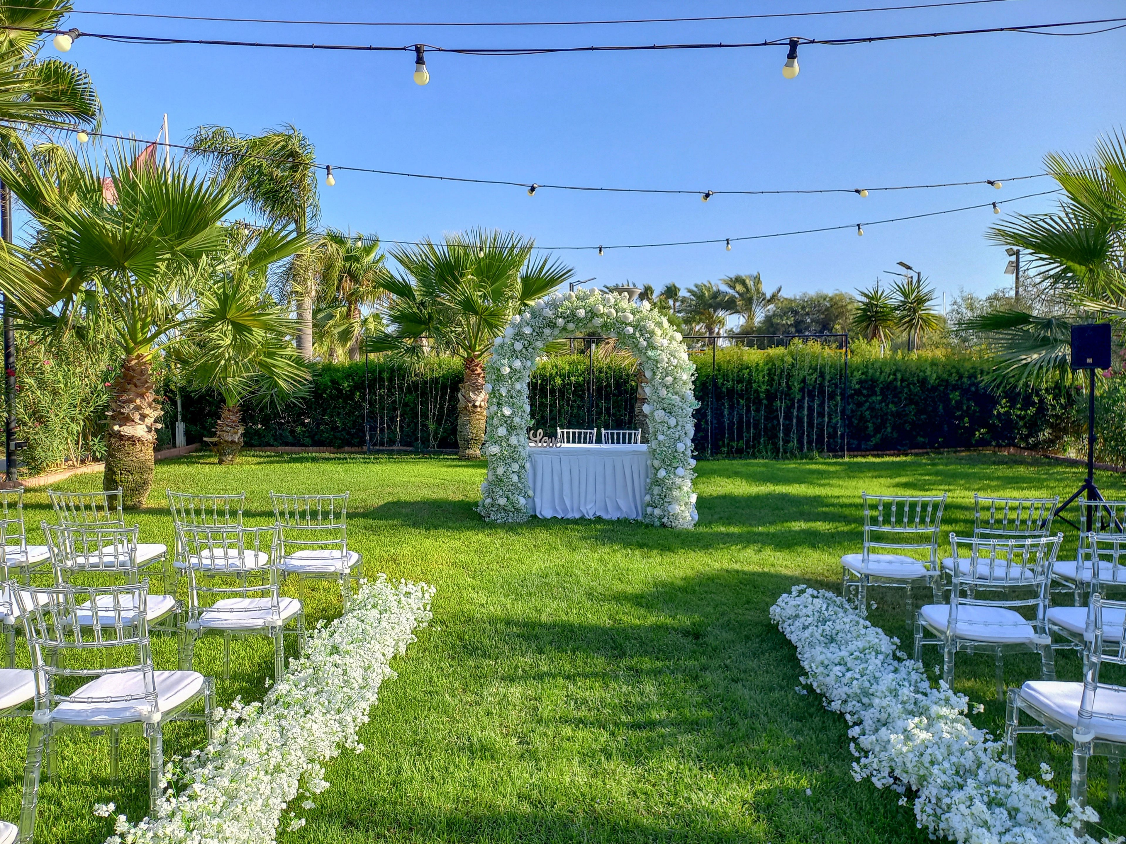 An outdoor wedding ceremony space is arranged on a lush green lawn with a white floral arch as the focal point. Transparent chairs are lined up in rows on both sides of a central aisle decorated with white flowers. String lights are suspended above the seating area, adding a festive touch to the open-air setting. Palm trees and dense greenery surround the venue, creating a serene and intimate atmosphere under a clear blue sky.
