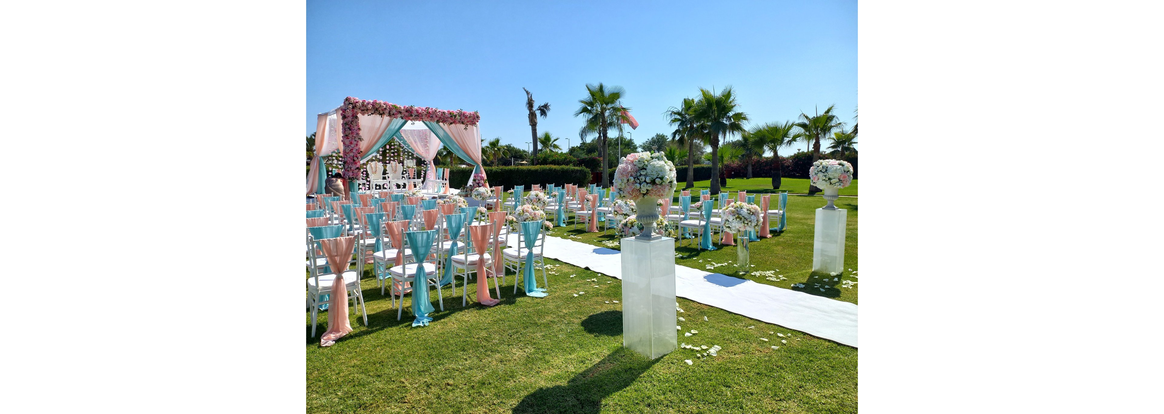 An elegant outdoor wedding ceremony is arranged on a manicured lawn under a clear blue sky. Rows of white chairs are decorated with pastel pink and turquoise fabric sashes facing a floral-draped mandap or arch. Tall glass stands hold large flower arrangements along a white aisle runner scattered with petals. Palm trees and distant greenery frame the romantic, tropical setting. The scene suggests a daytime celebration with a soft, pastel color palette.