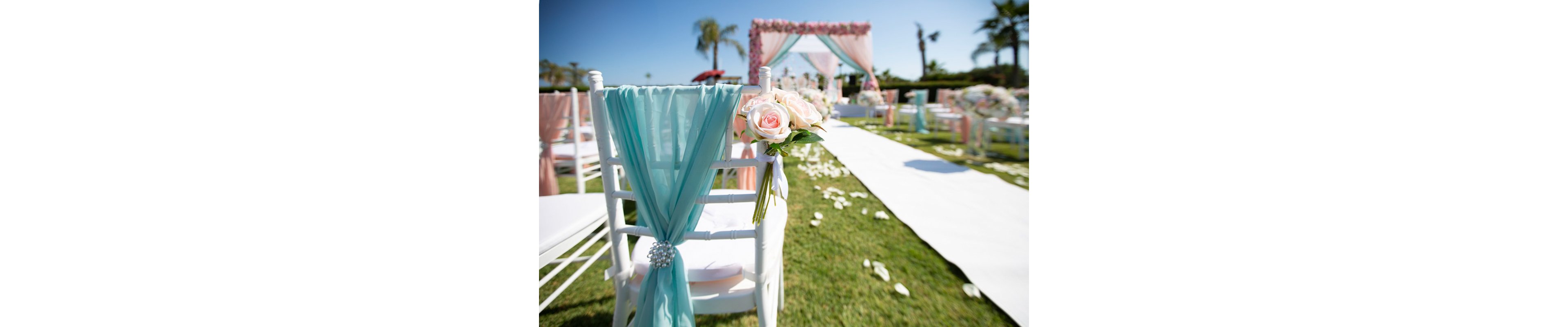 A white chair decorated with soft pink roses and turquoise fabric stands beside an outdoor wedding aisle. The scene features a long white runner leading to a floral arch in the distance. Petals are scattered on the grass, and additional chairs and flower arrangements line the pathway. Palm trees and a clear blue sky create a bright, romantic atmosphere.