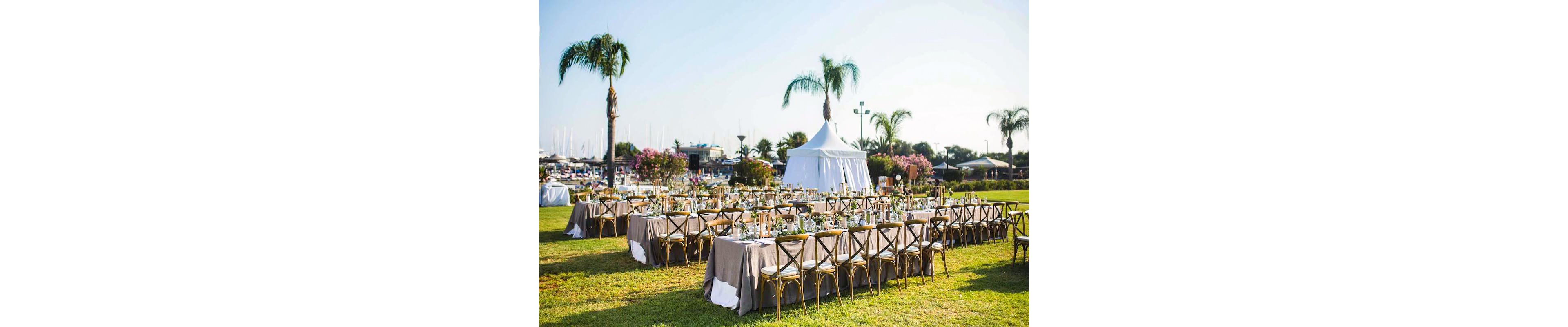 An elegant outdoor event setup features long banquet tables arranged on a grassy lawn. The tables are covered with neutral tablecloths and decorated with glassware, plates, and floral centerpieces. A white tent stands in the background surrounded by palm trees and flowering shrubs, suggesting a festive celebration or wedding reception. The scene is brightly lit with clear blue sky, creating a cheerful and inviting atmosphere near a marina.