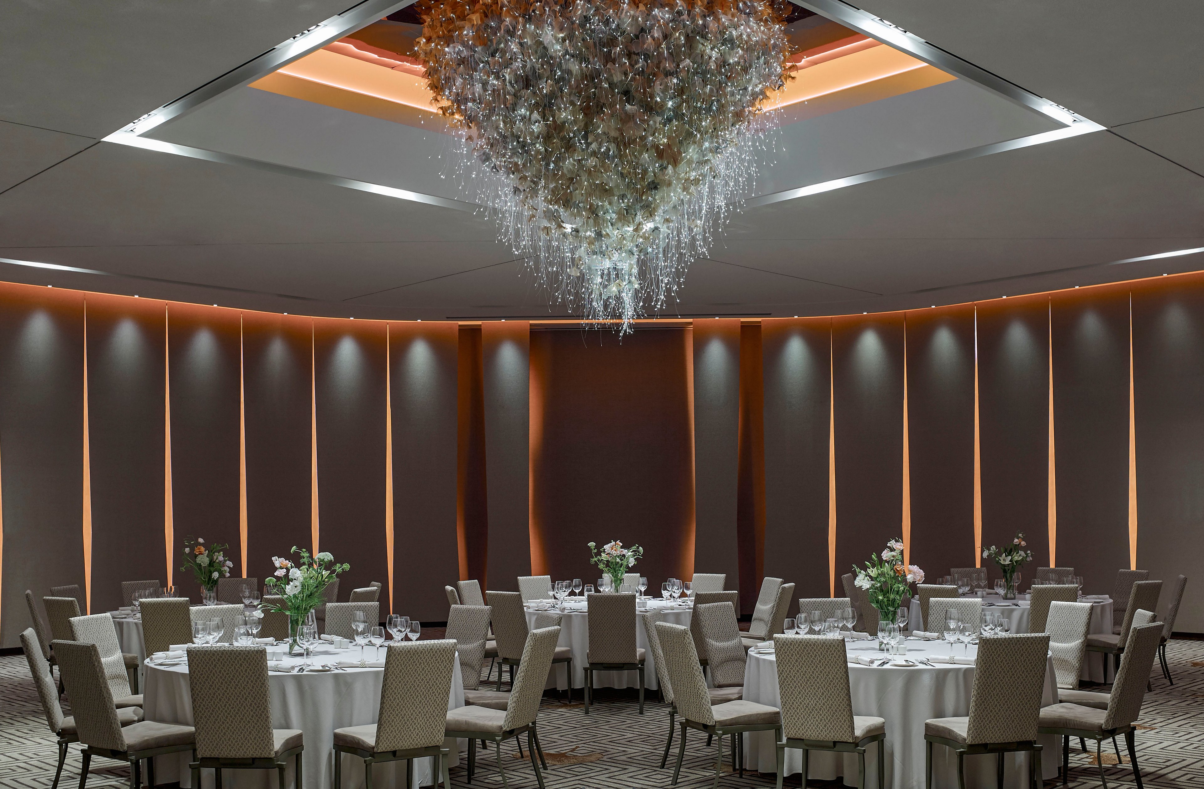 A sophisticated banquet hall is arranged with round tables covered in white linens and surrounded by beige upholstered chairs. Each table is decorated with floral centerpieces and glassware, ready for a formal event or reception. The room features tall vertical wall panels with warm accent lighting that creates a soft, inviting glow. A large, intricate crystal chandelier hangs from a recessed ceiling, serving as the dramatic focal point of the space.