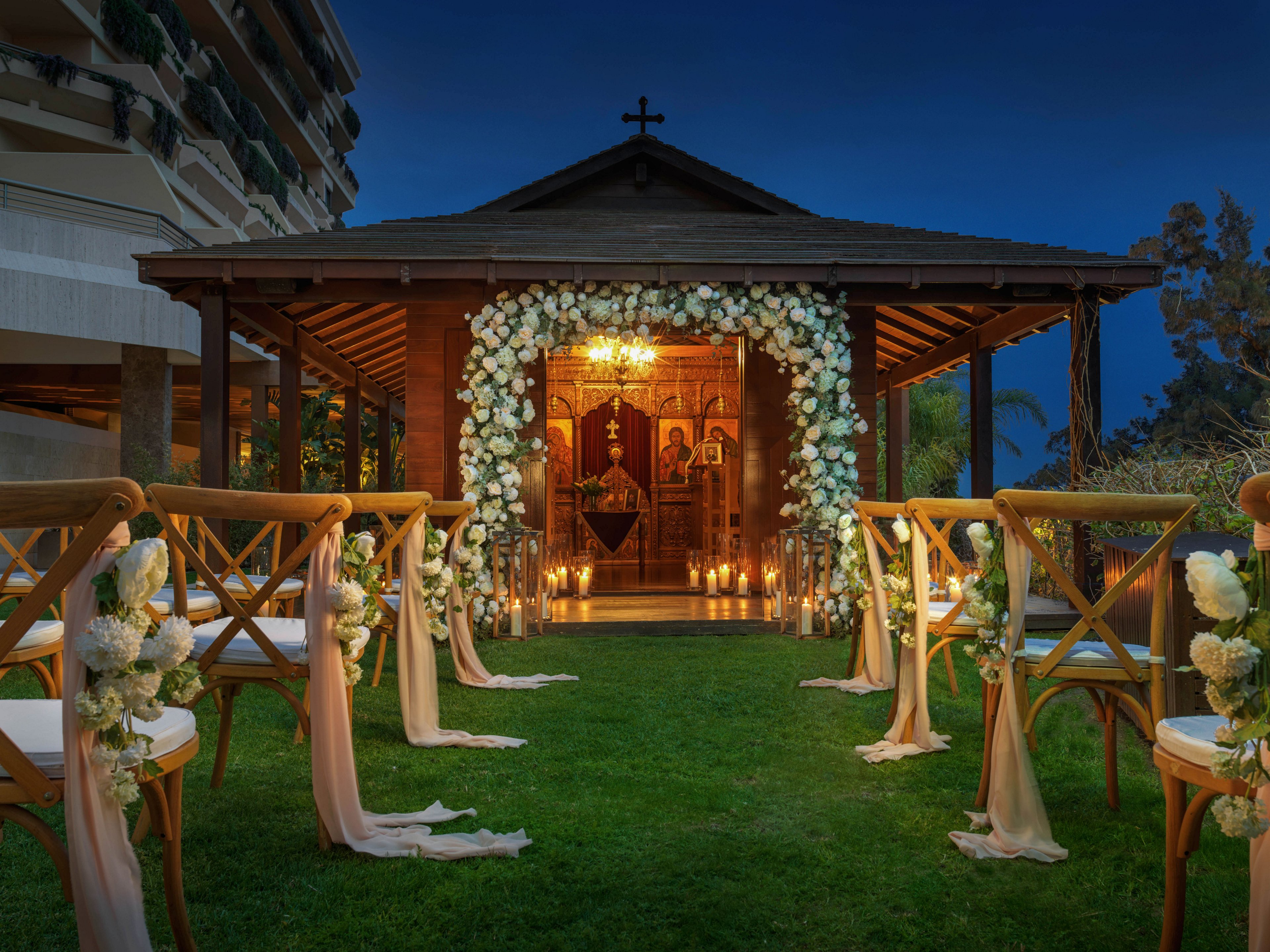 An outdoor wedding ceremony is set up in front of a small wooden chapel at dusk. Wooden chairs decorated with white flowers and flowing fabric line a grassy aisle leading to the entrance. The chapel doorway is framed with a lush floral arch and softly lit by warm candles inside. Religious icons and a cross on the roof emphasize the sacred setting, while the surrounding buildings and trees create a serene backdrop.