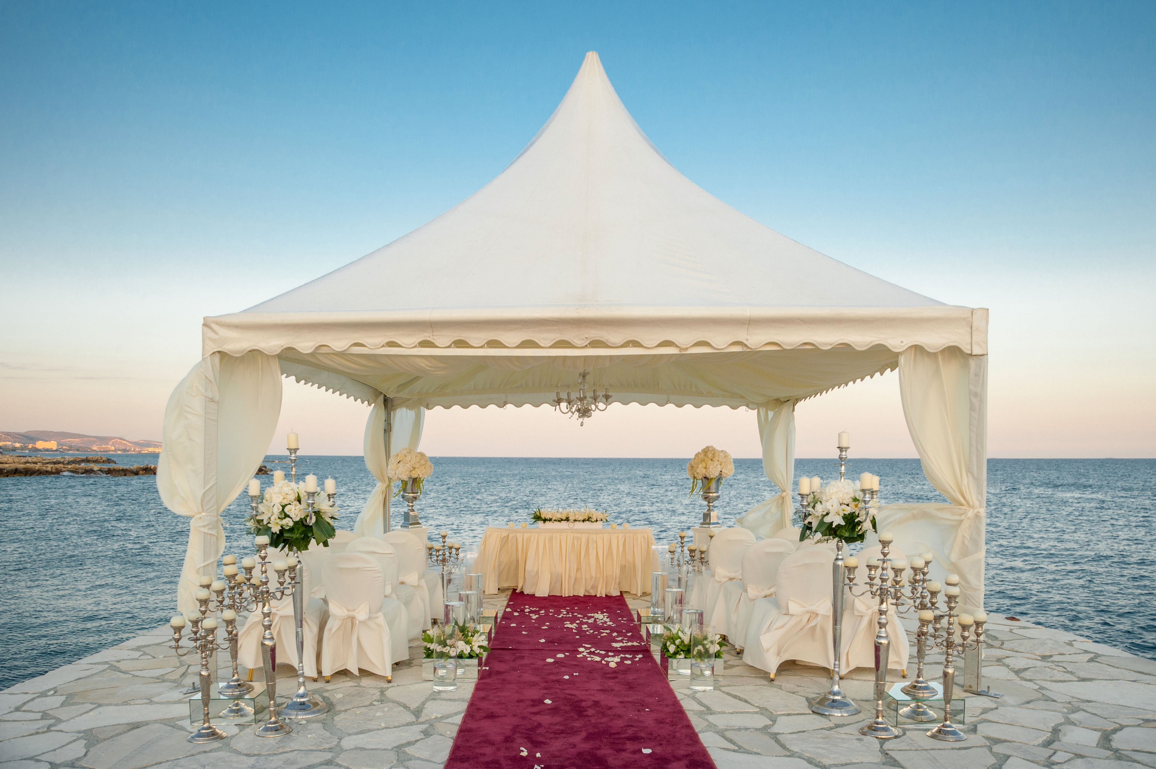 A white canopy tent is set up for a wedding ceremony by the sea, positioned on a stone platform extending over the water. Rows of white-covered chairs line a red carpet aisle leading to a decorated table with floral arrangements. Silver candle holders and vases add a touch of luxury, while white flowers and soft draping create a romantic atmosphere. The calm ocean and clear evening sky form a serene backdrop for the event.