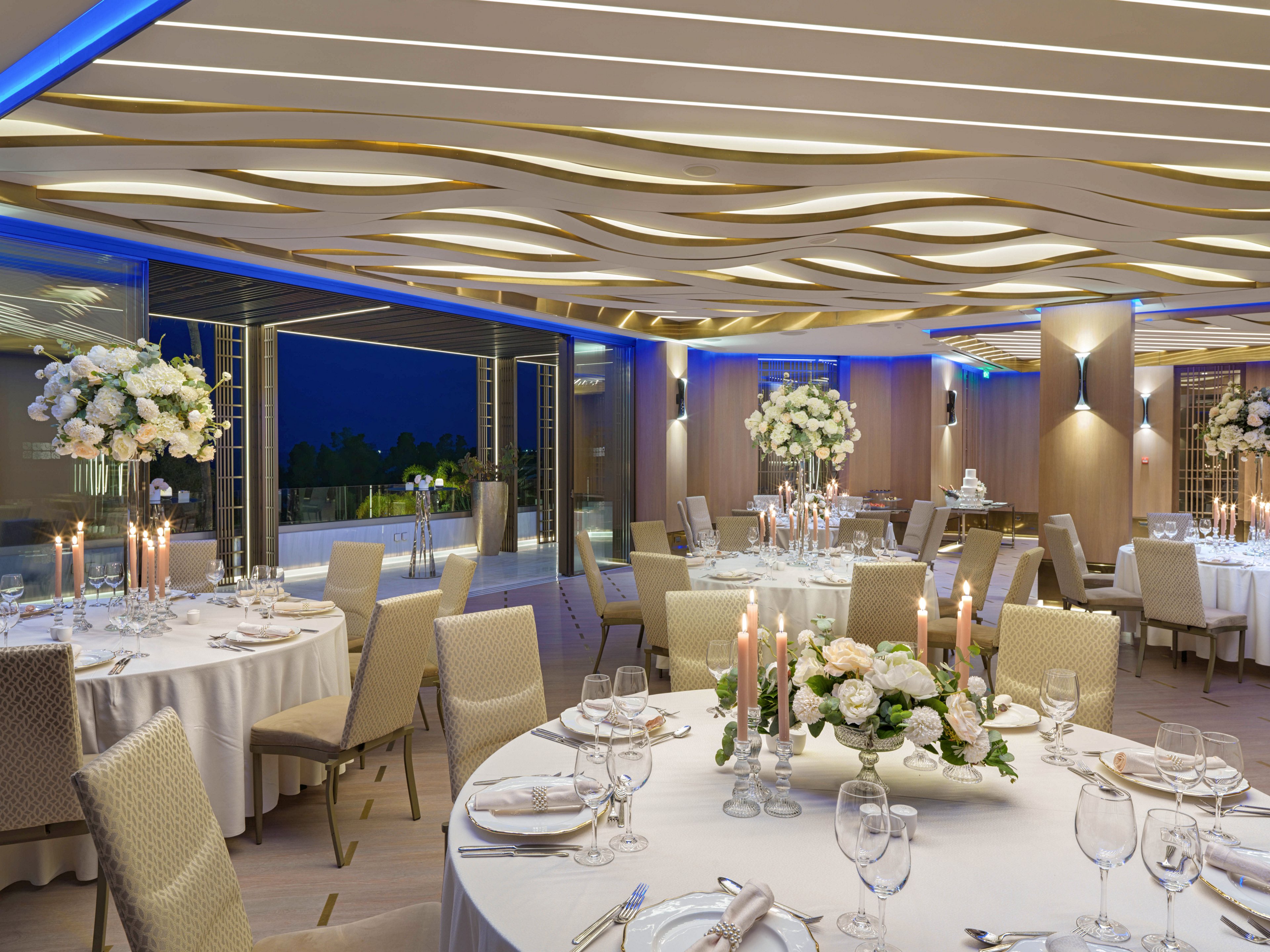 A sophisticated banquet hall is set for a formal event with round tables covered in white linens. Tall floral centerpieces with white flowers and greenery decorate each table, accompanied by lit taper candles in glass holders. The modern interior features a sculpted ceiling with wave-like patterns and blue accent lighting. Large windows open to an evening view, adding depth and ambiance to the scene.