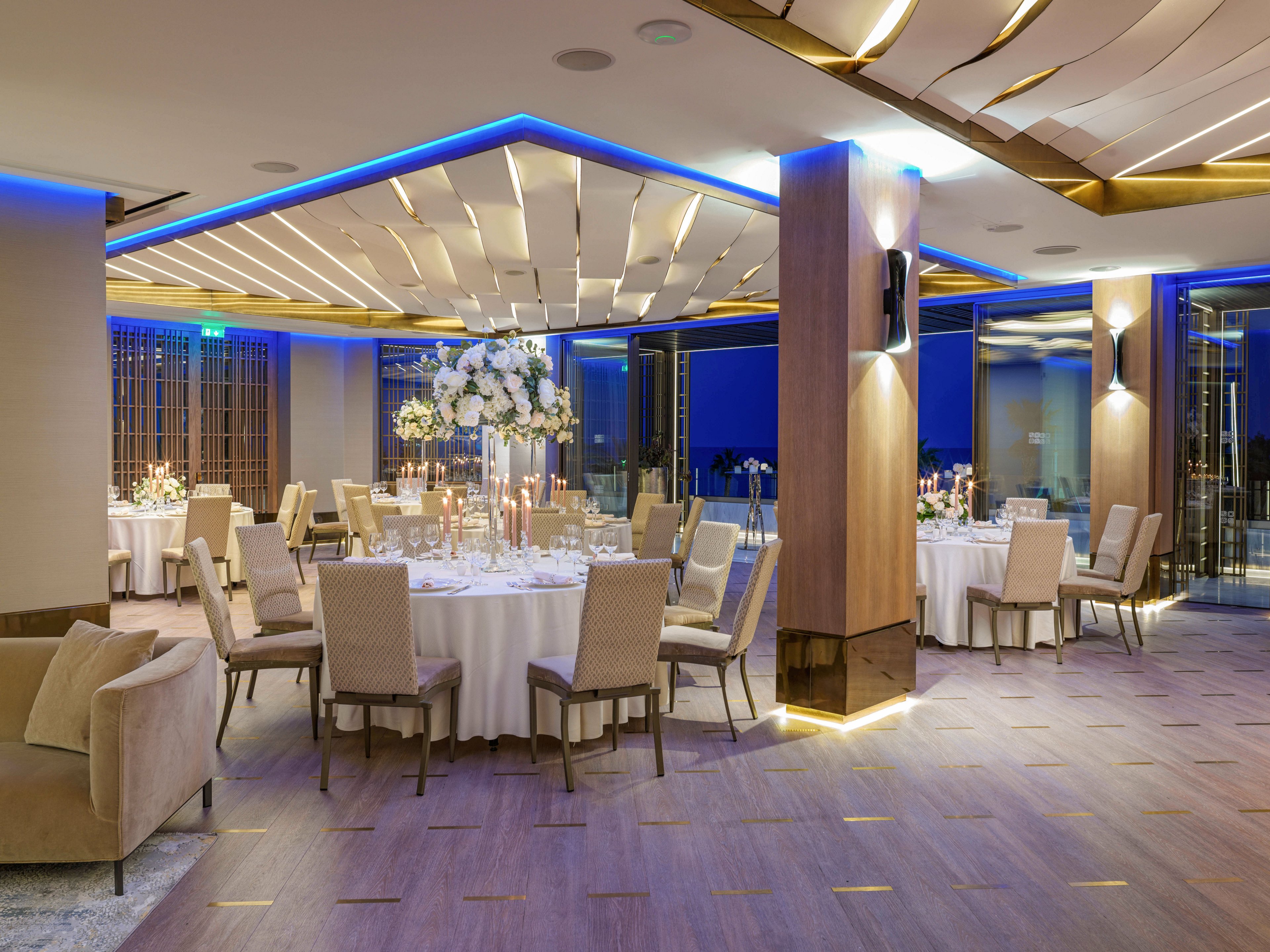 A sophisticated banquet hall is set up for a formal event with round tables covered in white linens. Neutral-toned upholstered chairs surround the tables, which are decorated with tall floral centerpieces and lit candles. The room features a modern ceiling design with blue accent lighting and warm recessed lights in the floor. Large windows reveal a night view outside, adding to the upscale ambiance of the space.