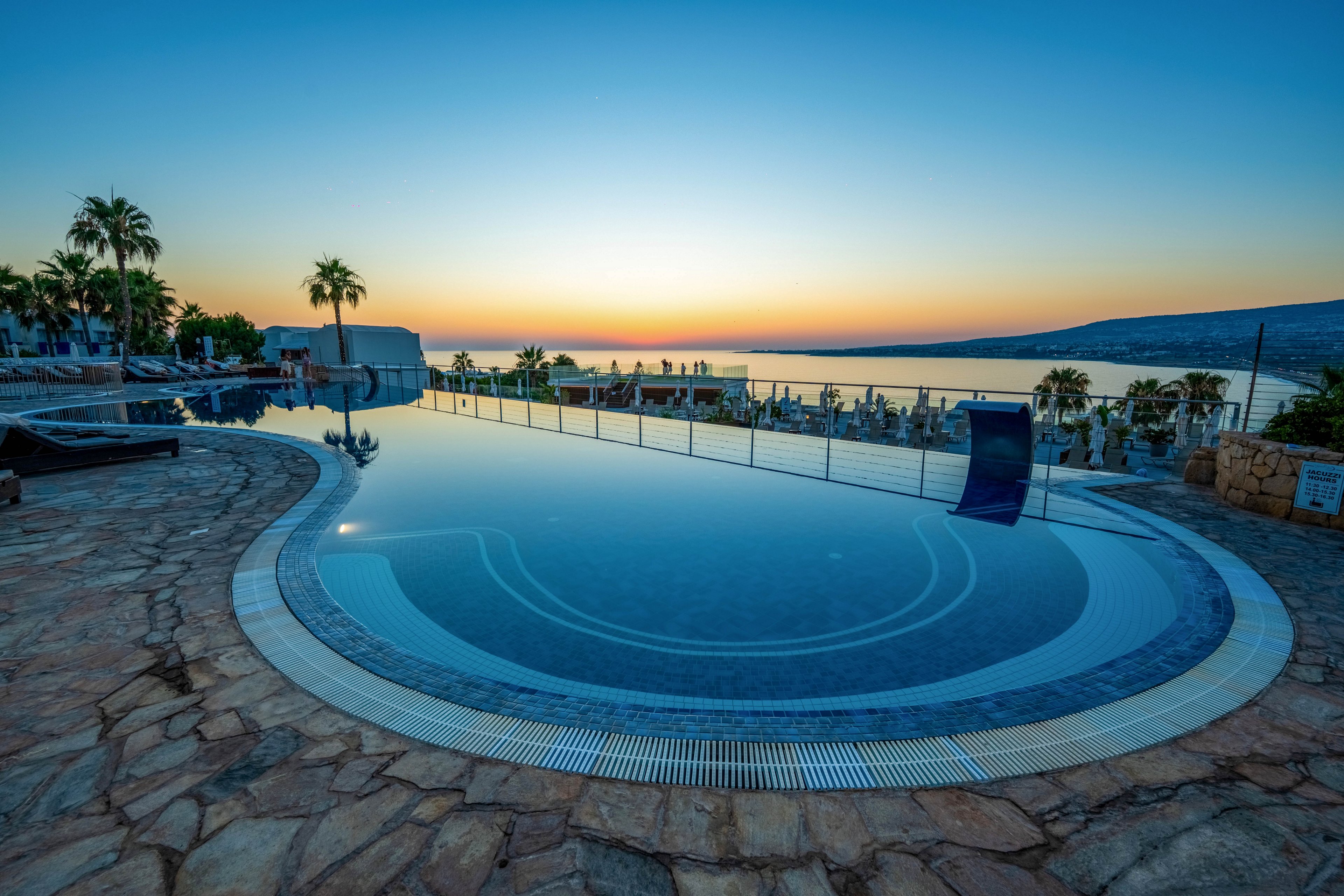 A curved infinity swimming pool overlooks a calm sea at sunset, with the sky fading from warm orange to deep blue. Palm trees, sun loungers, and umbrellas line the stone terrace around the pool, suggesting a relaxing vacation resort setting. Glass railings separate the pool area from the water below, enhancing the open, panoramic coastal view. The scene feels peaceful and luxurious, emphasizing leisure, serenity, and evening light by the ocean.