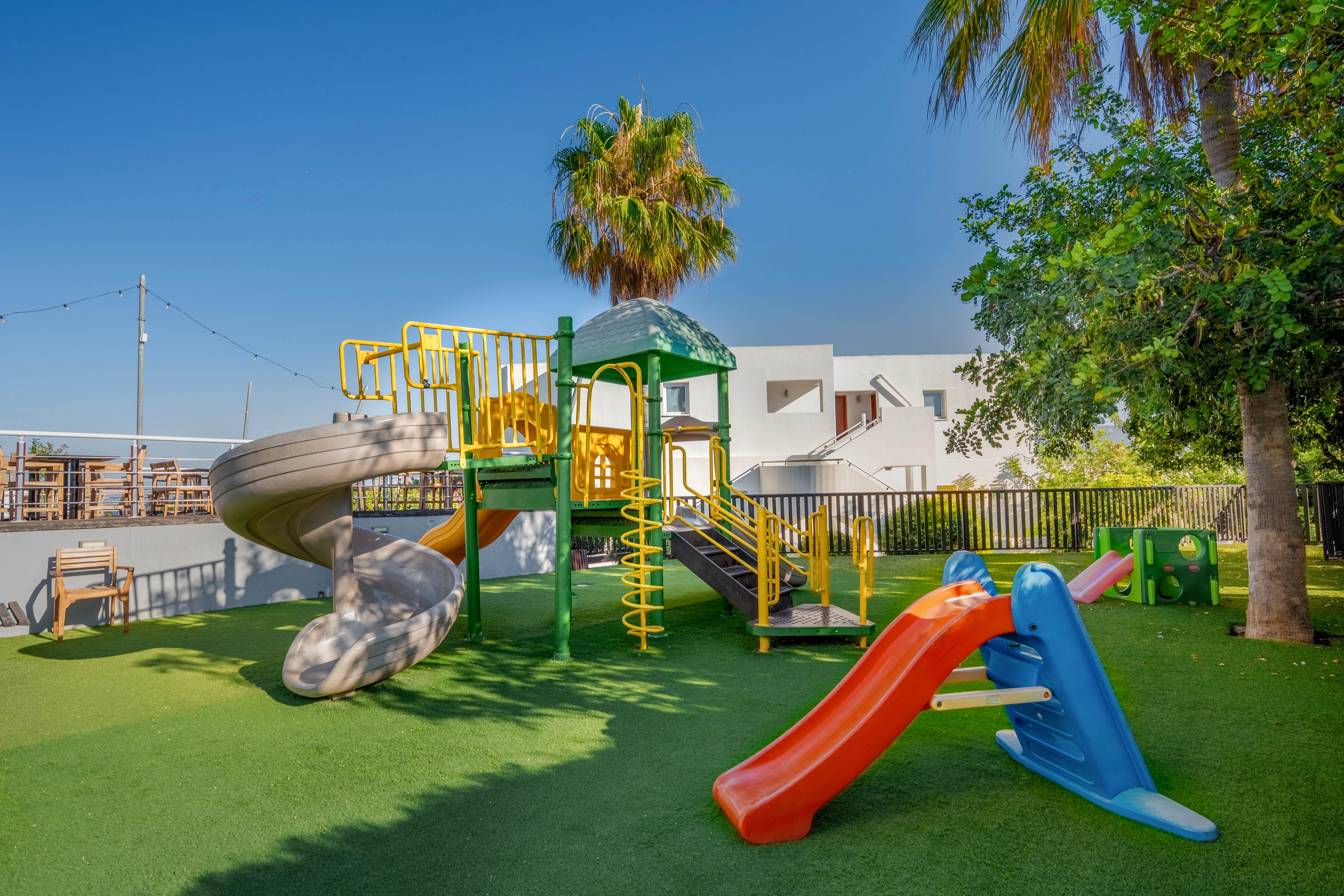 A bright outdoor playground features multiple slides, climbing structures, and ladders on a soft green surface. The equipment is painted in vivid colors including yellow, green, blue, orange, and red, creating a playful atmosphere. Palm trees and other greenery surround the play area, with modern white buildings visible in the background. The scene is lit by clear, sunny daylight, suggesting a warm and inviting environment for children.