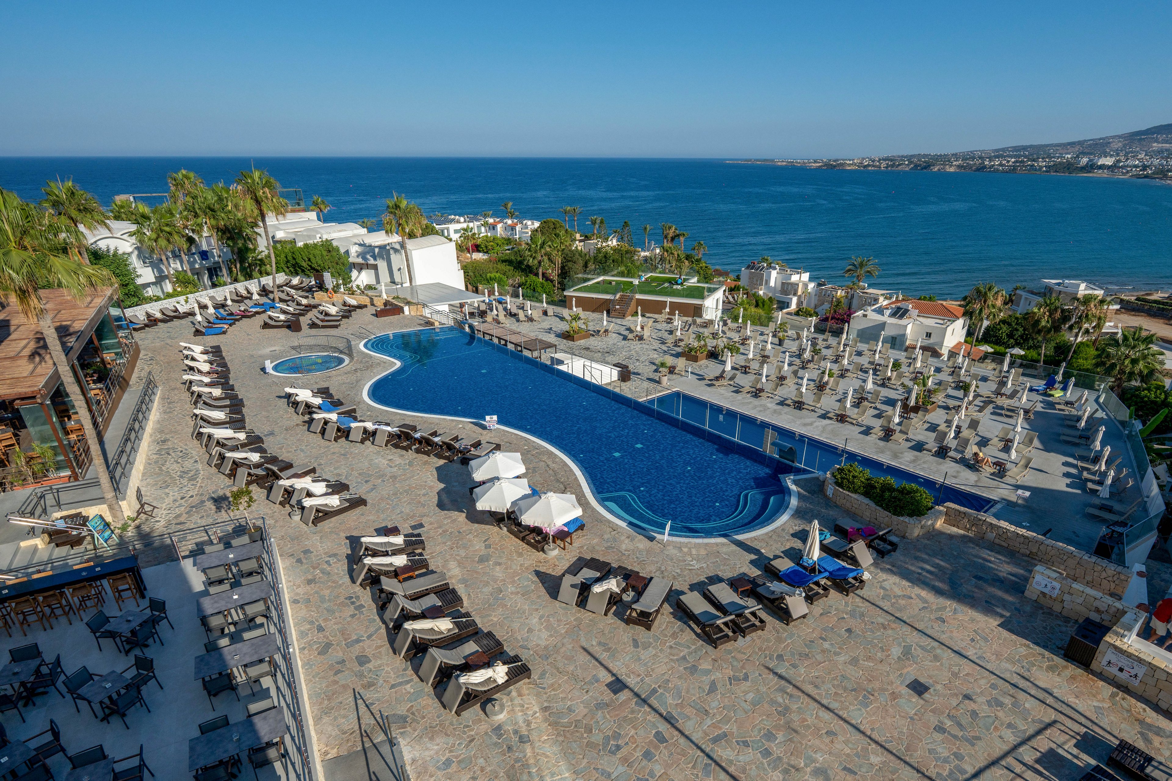 A large curved swimming pool sits at the center of a seaside resort terrace, surrounded by rows of sun loungers and umbrellas. The scene overlooks a calm blue sea and distant coastline under a clear sky. Palm trees and white buildings frame the pool area, while outdoor dining tables and chairs line the stone-paved terrace. The overall atmosphere is relaxed and inviting, suggesting a sunny vacation destination.