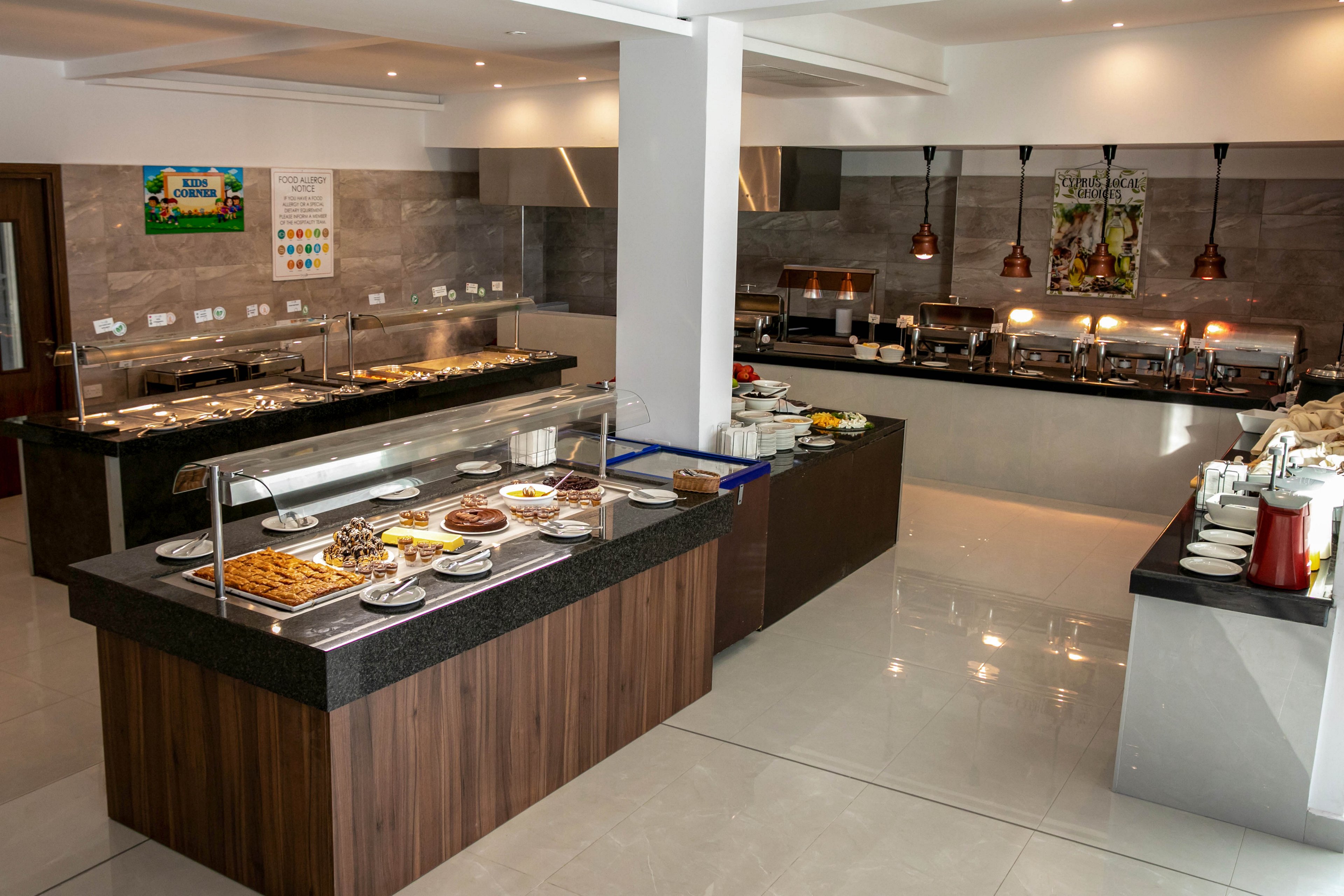 A spacious self-service buffet restaurant features multiple food islands with granite countertops and wooden bases. Various dishes and desserts are neatly arranged on plates and trays under glass sneeze guards. In the background, heated serving stations with copper lamps and stainless steel equipment line the wall. The room has bright, clean tile flooring and contemporary decor, creating a polished, inviting dining atmosphere.