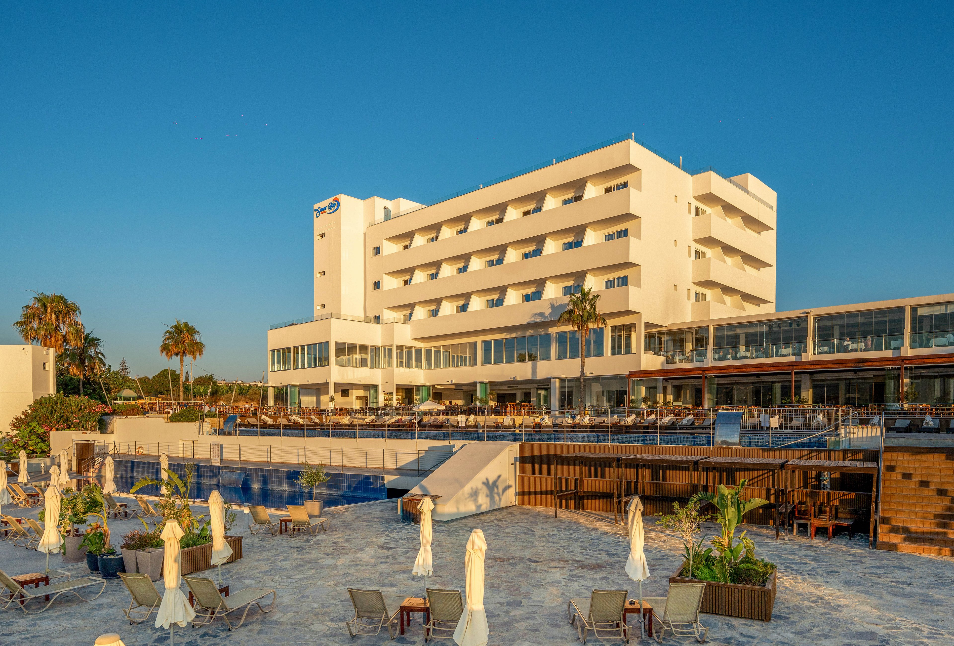 A large multi-story resort hotel stands beside a spacious beachfront terrace under warm sunset light. Sun loungers and closed umbrellas are arranged around an outdoor pool area, facing the sea. Palm trees and landscaped planters add a tropical feel to the scene. The clear blue sky and golden tones create a relaxed vacation atmosphere.