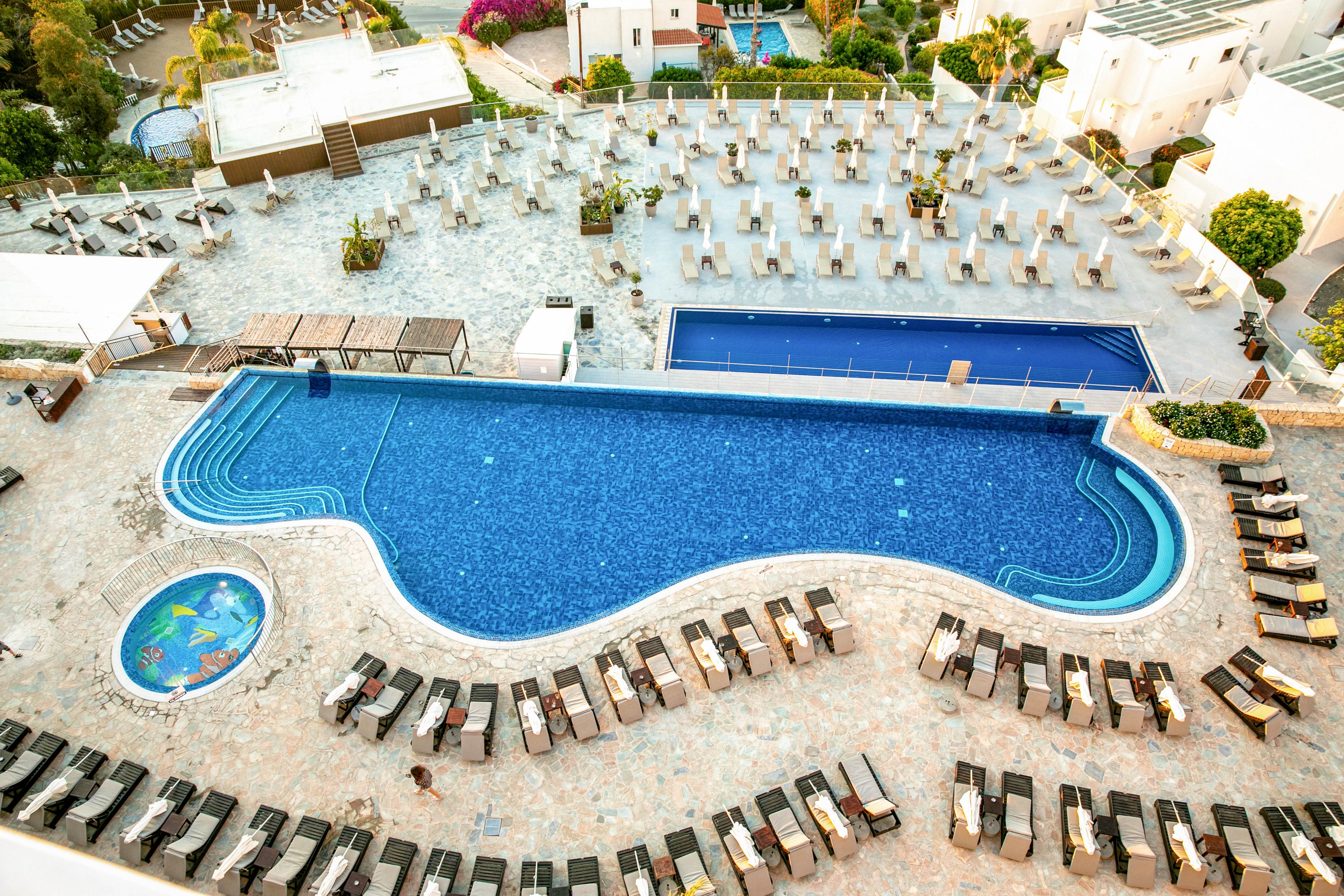A large, curving outdoor swimming pool is surrounded by numerous sun loungers neatly arranged on a tiled terrace. A smaller rectangular pool sits behind it, with additional rows of chairs and umbrellas creating a structured resort layout. In one corner, a colorful circular children’s pool adds a playful element to the scene. Warm lighting and bright colors suggest a sunny vacation atmosphere at a Mediterranean-style hotel.