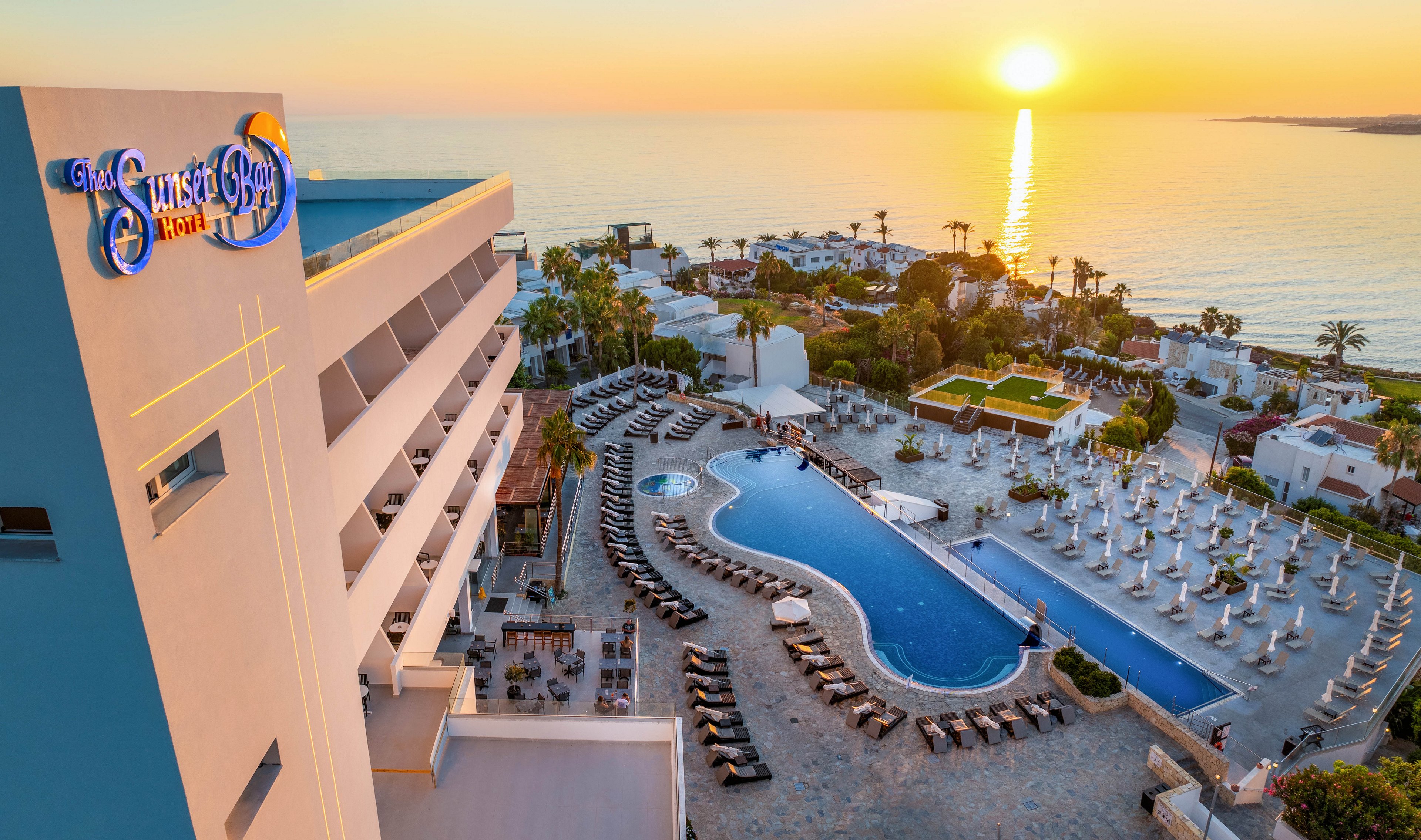 A large seaside resort is captured from above at sunset, overlooking an expansive pool area and rows of sun loungers. The hotel building stands on the left, with balconies facing the ocean and a colorful sign near the rooftop. The pool curves through the terrace, surrounded by neatly arranged umbrellas and palm trees. In the background, the sun sets over calm water, casting a golden reflection across the sea and warm light over the entire scene.