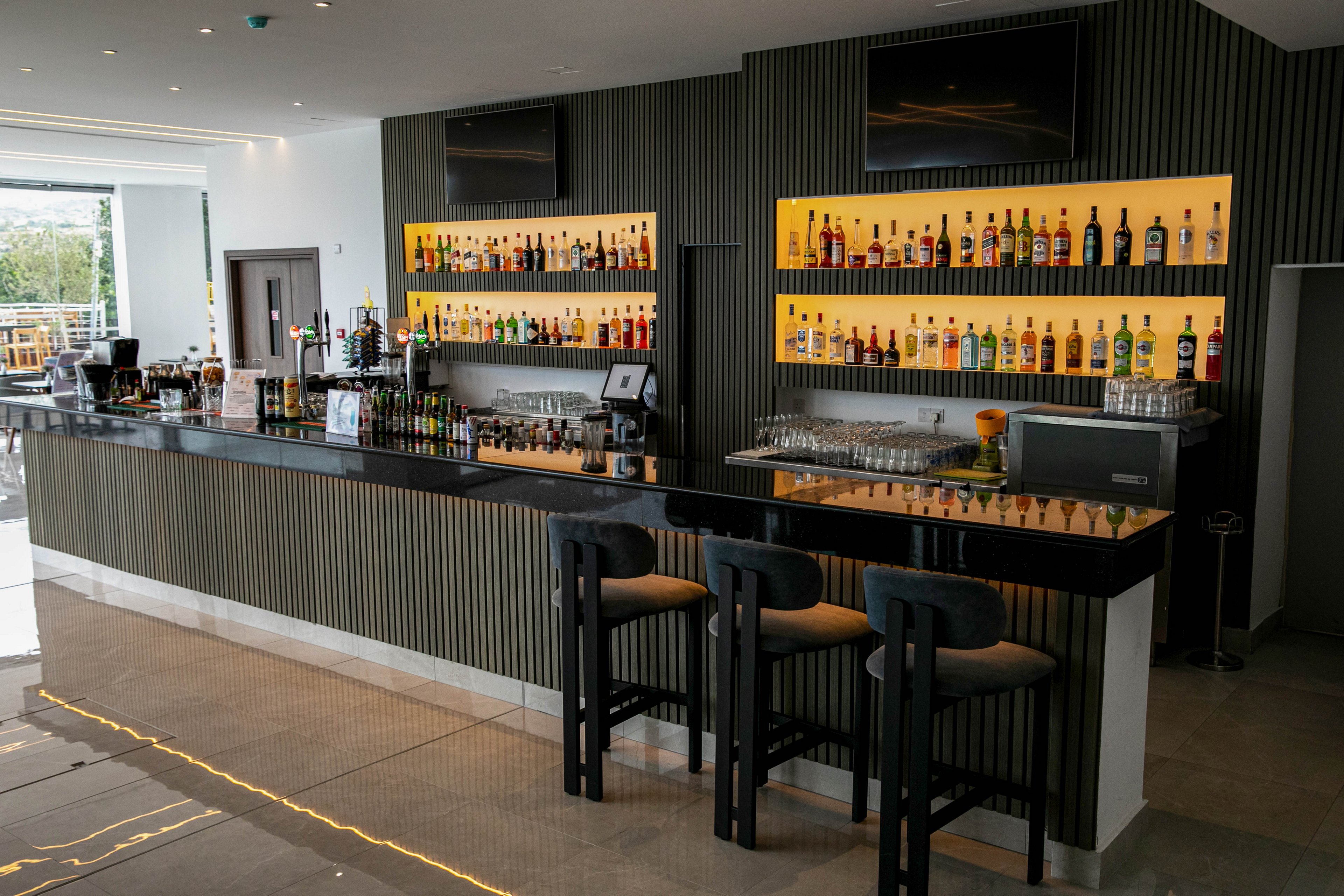 A sleek contemporary bar interior features a long black countertop with three high stools in the foreground. Backlit shelves display a wide variety of liquor bottles arranged in rows behind the bar. Glassware and bar equipment are neatly organized along the counter, creating a clean and professional look. Large windows on the side bring in natural light, enhancing the modern, upscale atmosphere.