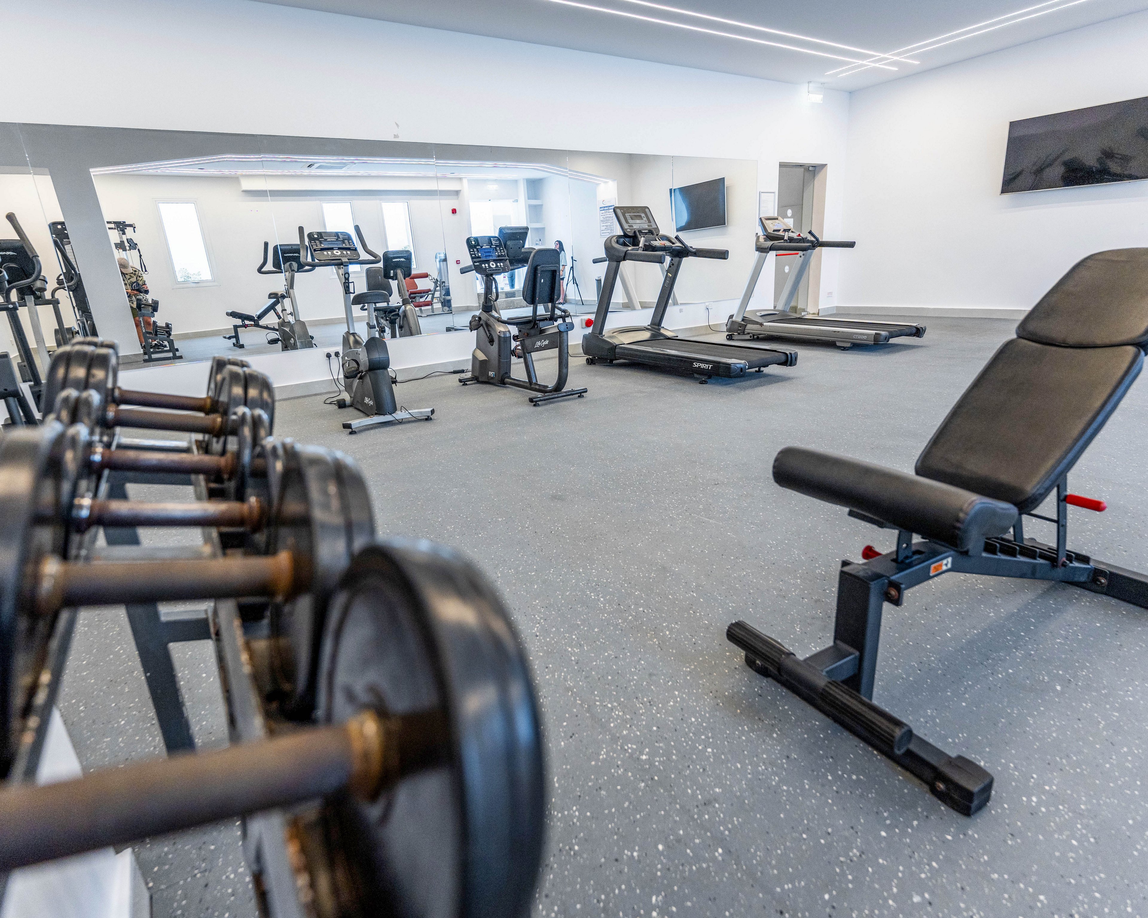 A bright, modern gym interior features free weights in the foreground and various cardio machines such as treadmills and stationary bikes in the background. A padded weight bench sits to the right on a speckled rubber floor, with large mirrors reflecting the equipment and expanding the space. The room is clean and minimal, with white walls, ceiling lights, and a wall-mounted television adding to the contemporary feel. The overall setting suggests a well-equipped fitness center designed for strength training and cardio workouts.