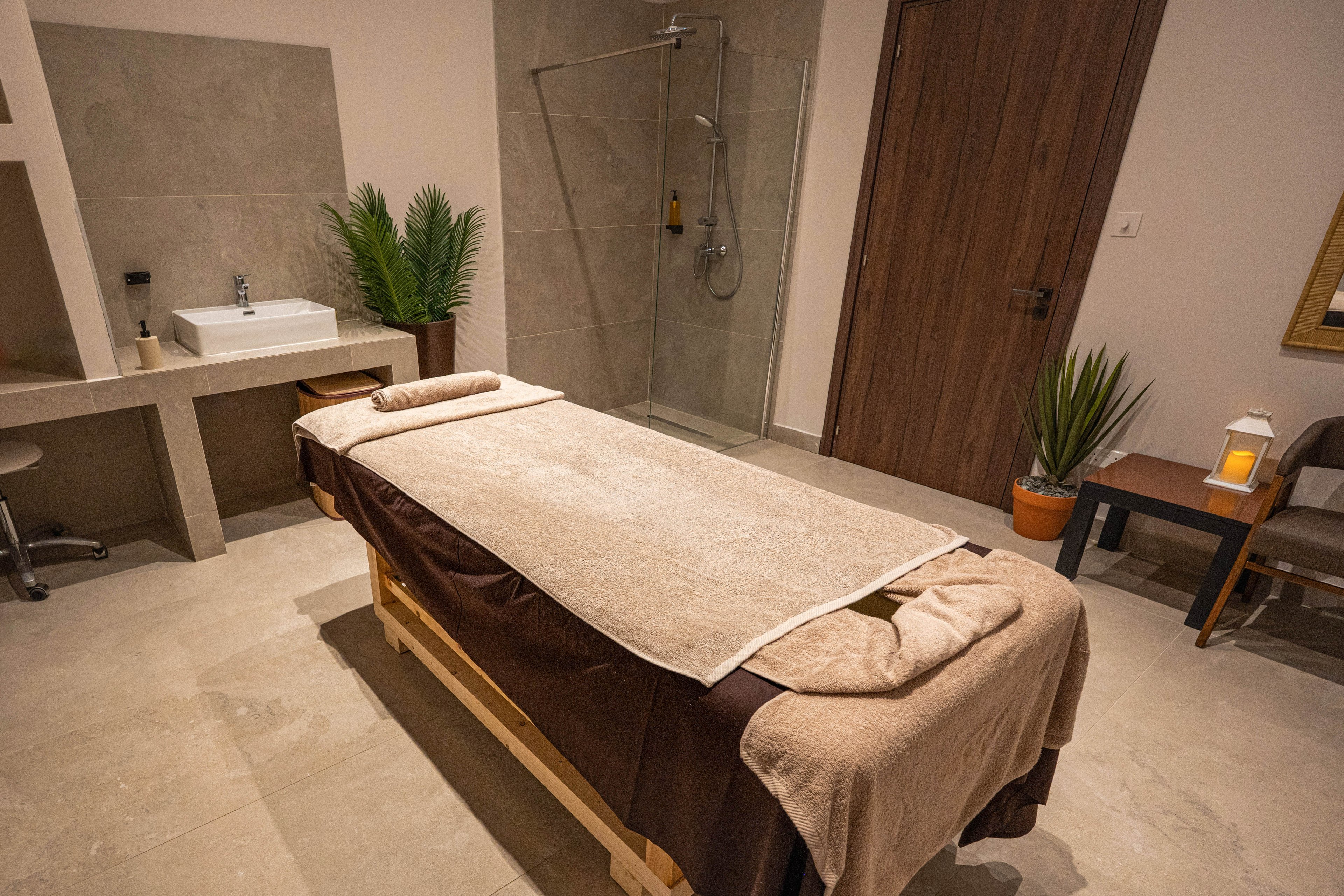 A neatly prepared massage table covered with beige towels stands in the center of a modern spa treatment room. The space features neutral stone flooring, a walk-in shower, and a minimalist sink area. Potted green plants and a small lantern on a side table add warmth and relaxation to the setting. The overall atmosphere is calm, clean, and inviting for wellness treatments.