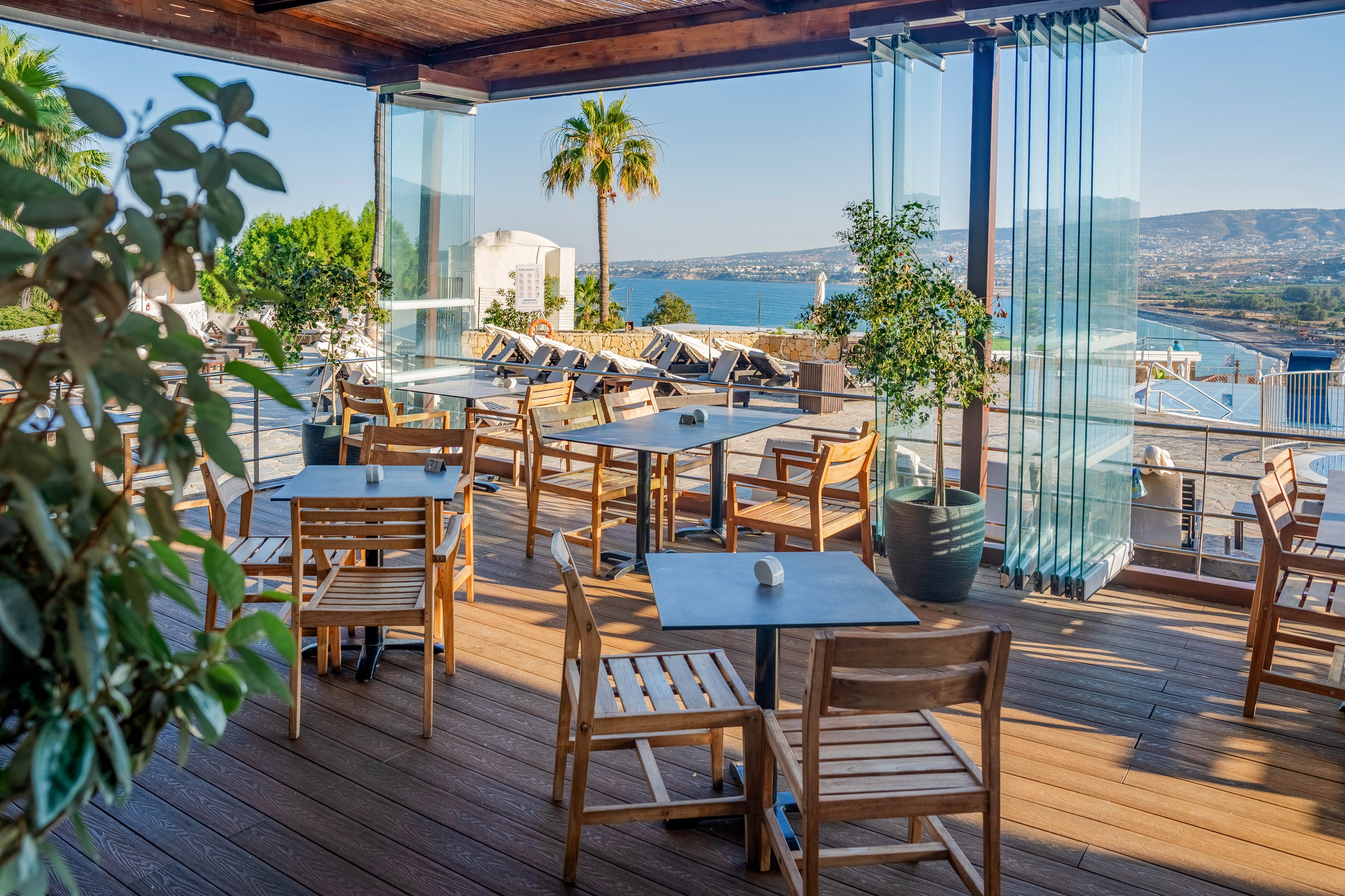 An open-air restaurant terrace features wooden tables and chairs arranged on a deck. Glass panels frame the space, offering clear views of a blue bay and distant hills. Sun loungers line a poolside area in the background, suggesting a resort setting. Lush green plants and a tall palm tree add a relaxed, Mediterranean atmosphere under bright daylight.