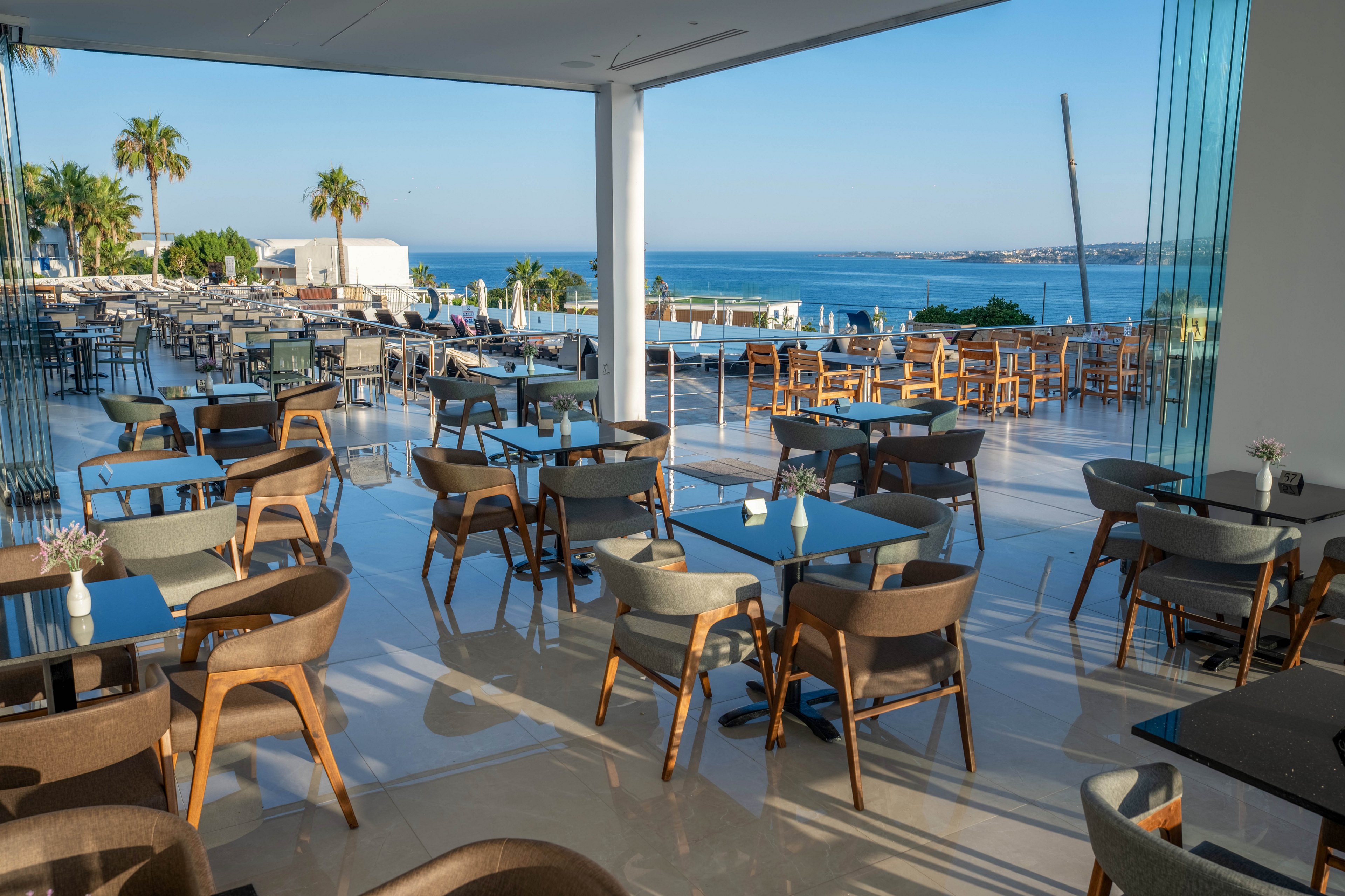 A contemporary open-air restaurant terrace overlooks a calm blue sea and distant coastline. Rows of wooden chairs and dark tabletops are neatly arranged on a glossy floor, with glass railings framing the view. Palm trees and sunlit buildings appear in the background under a clear sky, suggesting a warm coastal destination. The scene feels tranquil and inviting, ideal for relaxed outdoor dining with ocean views.