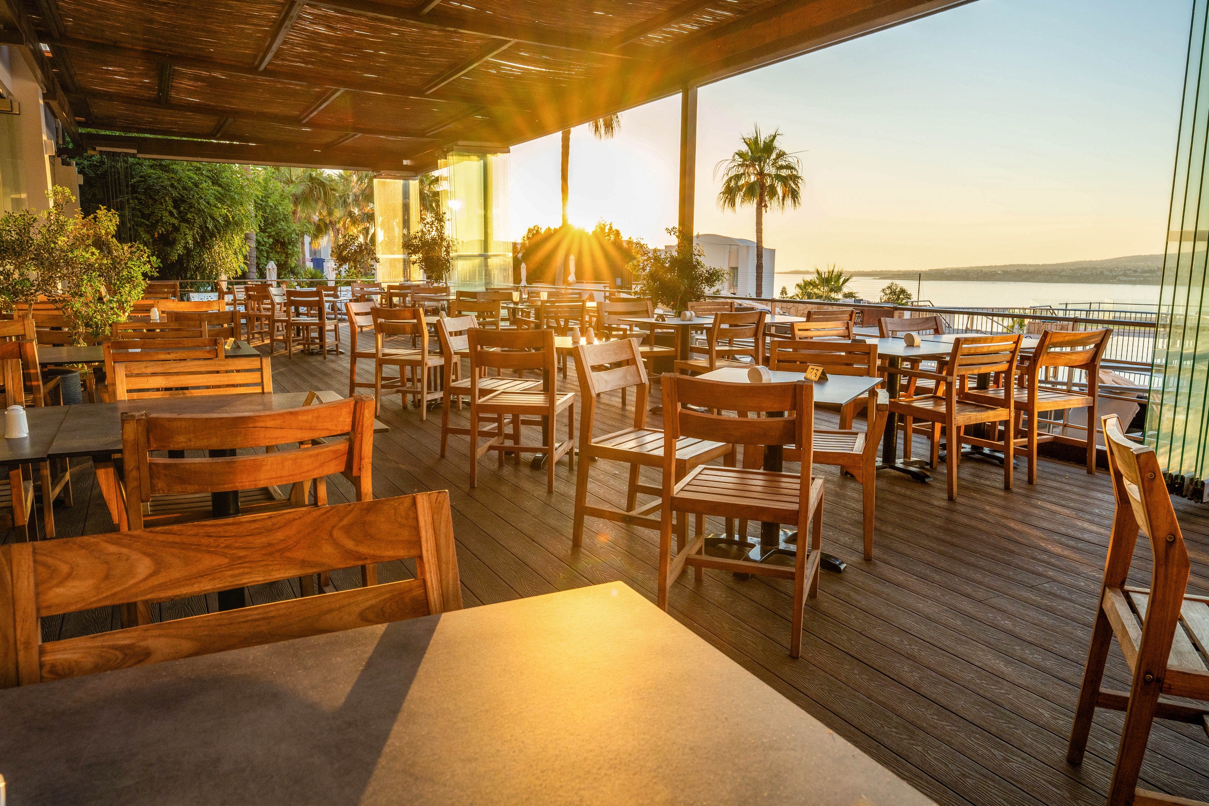 An inviting outdoor restaurant terrace is filled with wooden tables and chairs under a partial roof. Warm sunlight streams through the space, casting long shadows and highlighting the natural wood tones. Lush greenery and a tall palm tree frame the view toward a calm body of water in the distance. The scene feels peaceful and relaxed, suggesting a pleasant dining experience at golden hour.