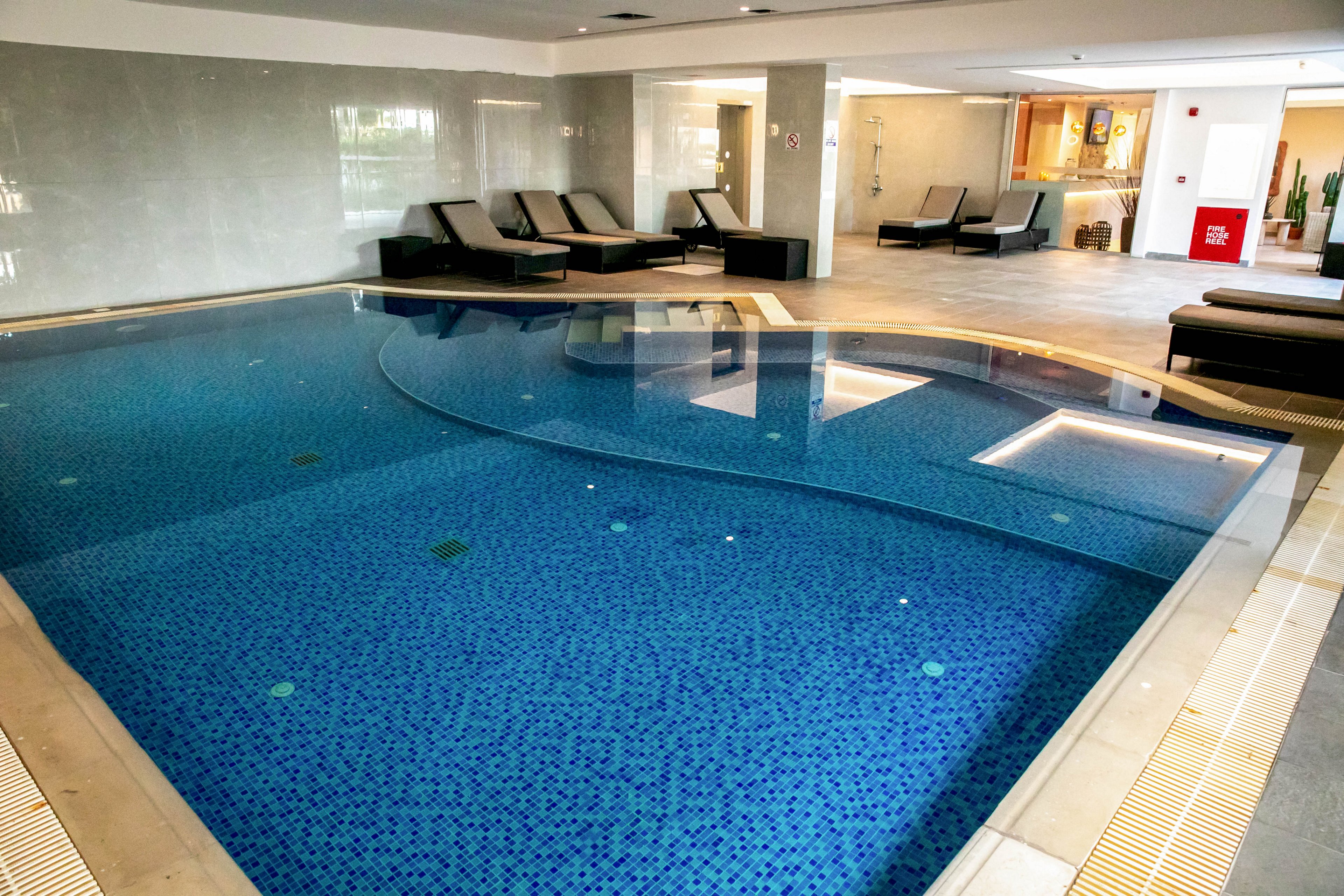 An indoor spa pool with blue mosaic tiles is the main focus of the image. Several dark cushioned loungers line the far side of the pool in a modern, minimalist setting. The room features light-colored walls, recessed lighting, and a clean, contemporary design. In the background, a small seating area and decor elements add to the relaxing atmosphere.