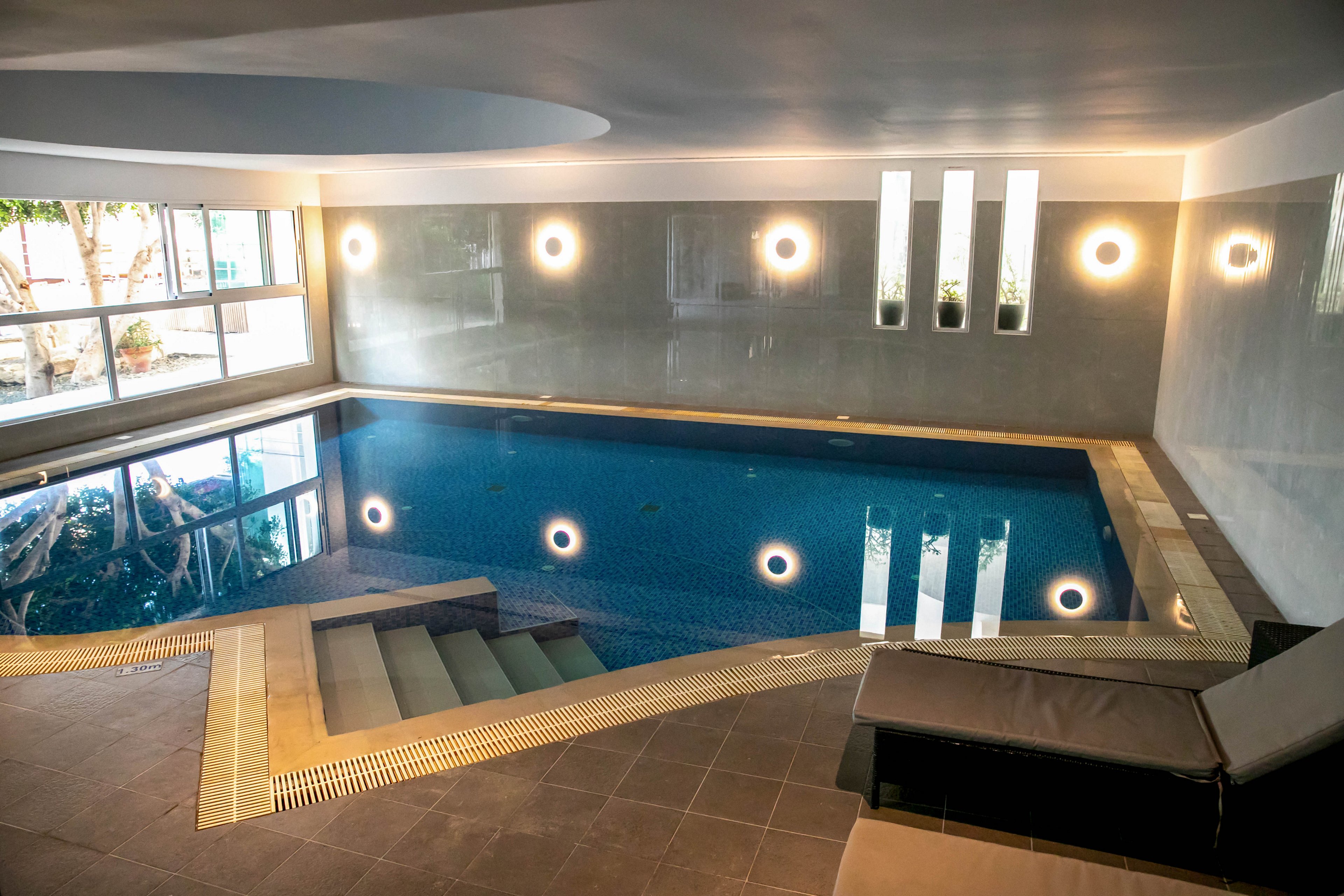 A contemporary indoor swimming pool is shown with clean lines and tiled steps leading into the water. Soft wall lights and vertical window panels create a calm, reflective atmosphere. A cushioned lounge chair sits in the foreground on the tiled deck, inviting relaxation. Large windows on the side wall allow natural light to brighten the serene pool area.