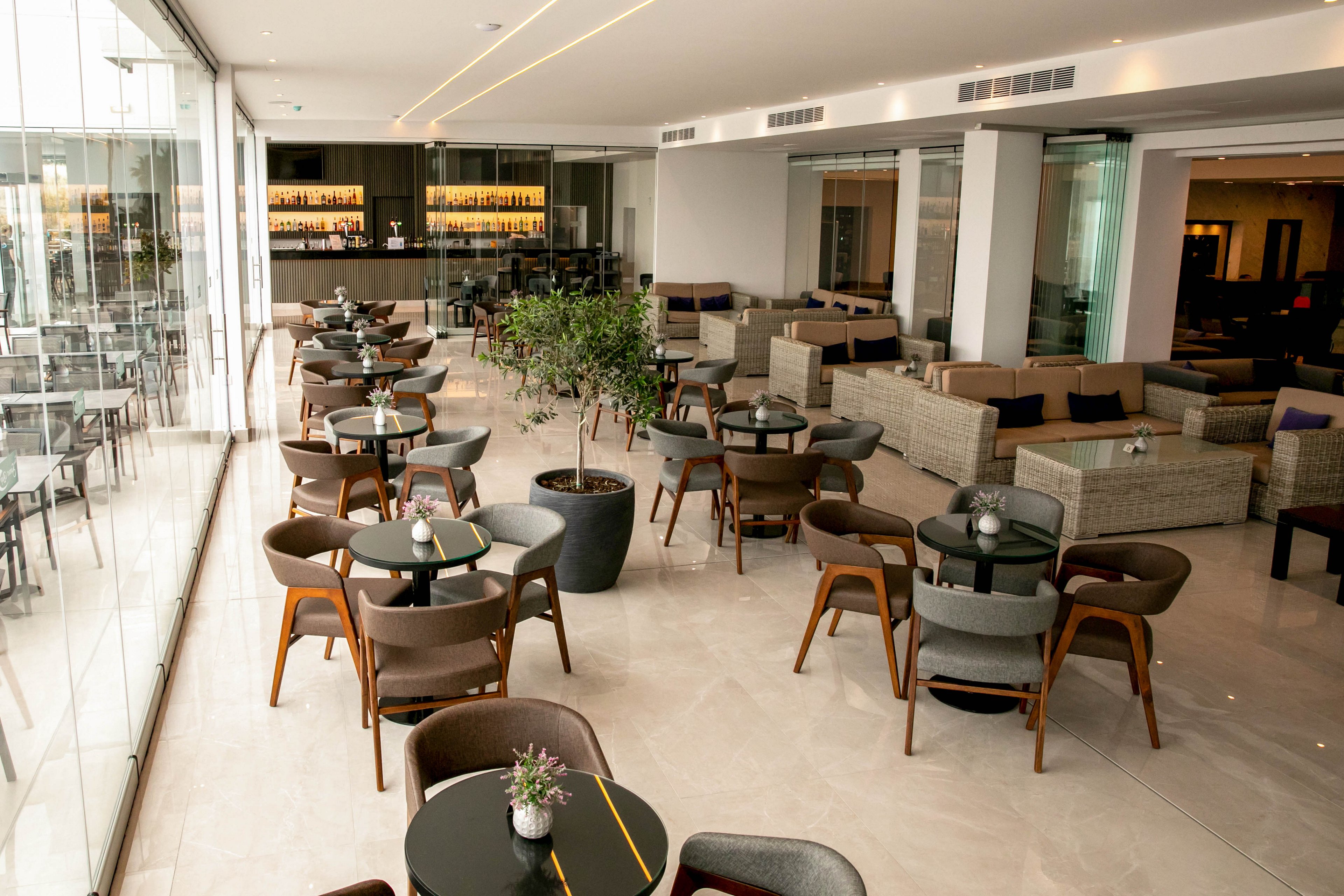Spacious contemporary lounge bar interior with multiple round tables and cushioned chairs arranged neatly across a polished floor. Large floor-to-ceiling glass windows line one side, allowing natural light to fill the room and reveal an outdoor view. A well-lit bar with shelves of bottles is visible at the back, complemented by sofa seating areas and minimalist decor. The overall atmosphere is clean, modern, and inviting, suitable for socializing or relaxing.