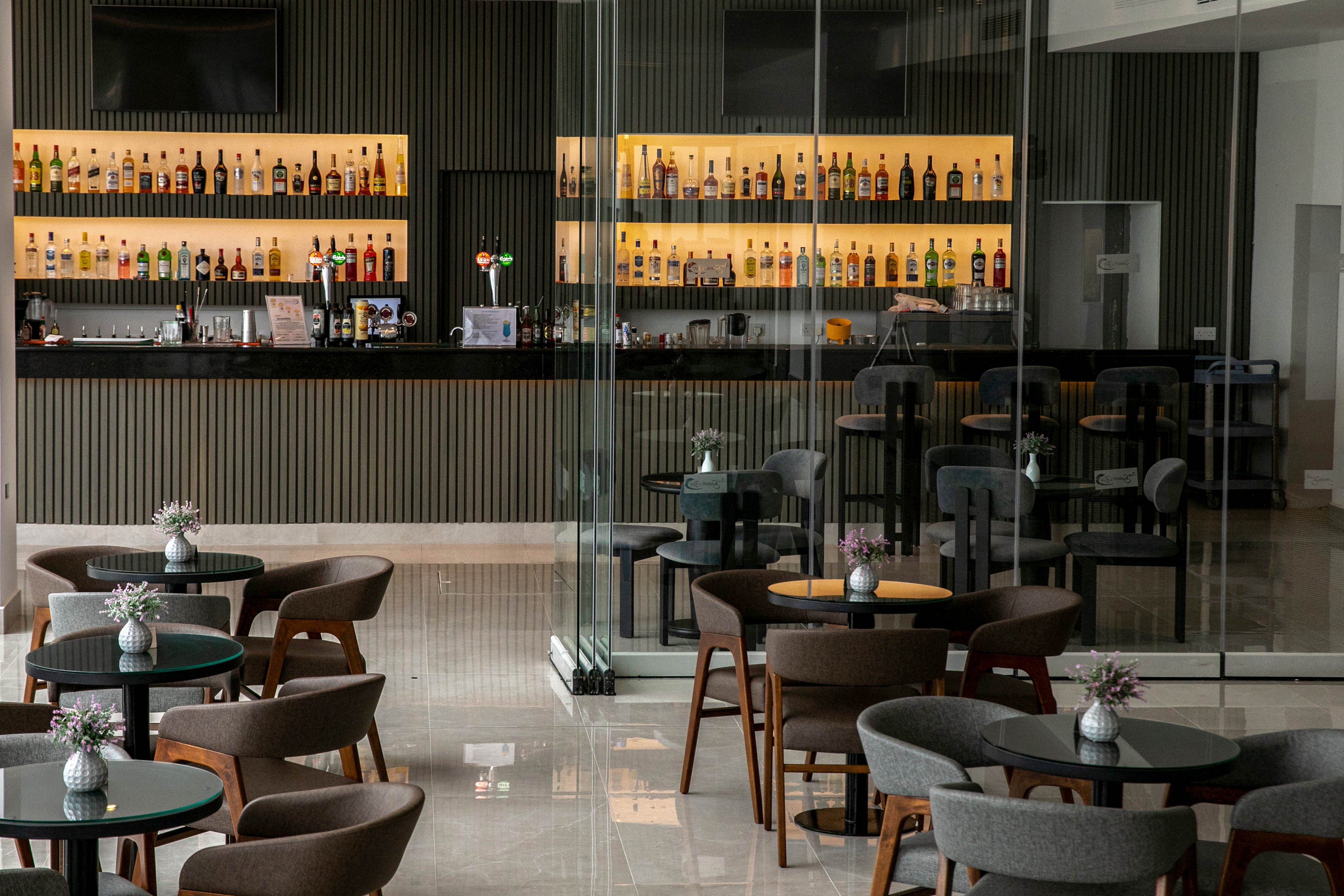 A contemporary bar interior features rows of liquor bottles displayed on backlit shelves behind a long counter. In the foreground, empty round tables with cushioned chairs are arranged neatly on a polished floor. Small vases with flowers decorate each table, adding a soft touch to the minimalist design. Glass partitions and clean lines create an open, sophisticated atmosphere suitable for hotel guests or business travelers.