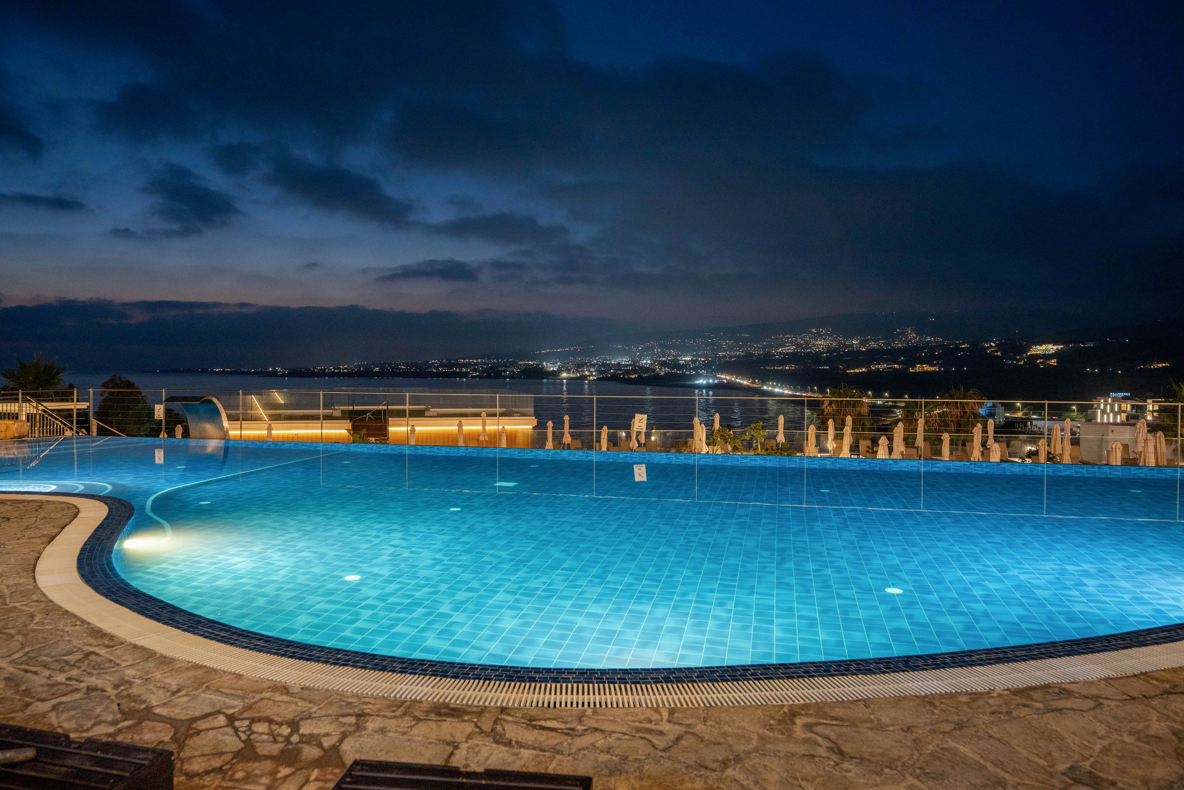 A glowing blue swimming pool is captured at night, with calm water reflecting the surrounding lights. The pool sits on a stone terrace, bordered by a glass railing that opens to a sweeping view of a coastal city. Distant urban lights trace the shoreline and hills under a dramatic cloudy sky. The scene evokes a tranquil, luxurious atmosphere with strong contrasts between the bright pool and dark horizon.