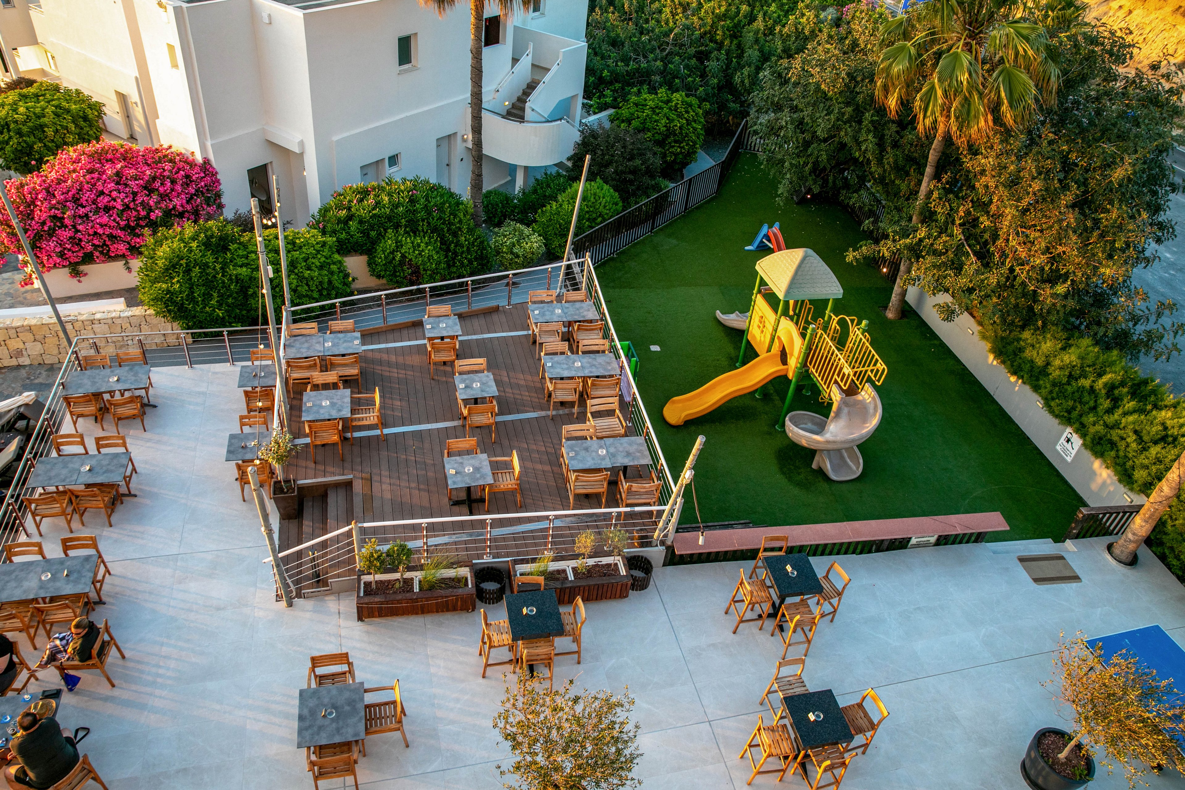 An elevated view shows an outdoor terrace cafe with multiple wooden tables and chairs neatly arranged on different levels. Adjacent to the seating area is a small playground featuring a bright green play structure with yellow slide and silver tube slide on artificial grass. Lush greenery, palm trees, and vibrant flowering bushes surround the space, creating a relaxed vacation atmosphere. Warm sunlight casts long shadows across the terrace, emphasizing the inviting, open-air setting.