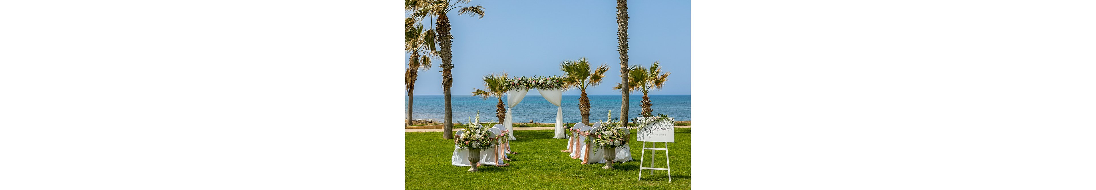 An elegant wedding ceremony setup is arranged on a lush green lawn overlooking a calm blue sea. A floral arch with white drapes stands at the front, framed by palm trees and facing rows of decorated white chairs. Large flower arrangements in urns line the aisle, featuring soft pastel blooms and greenery. A white welcome sign on an easel stands to the side, enhancing the romantic coastal atmosphere.