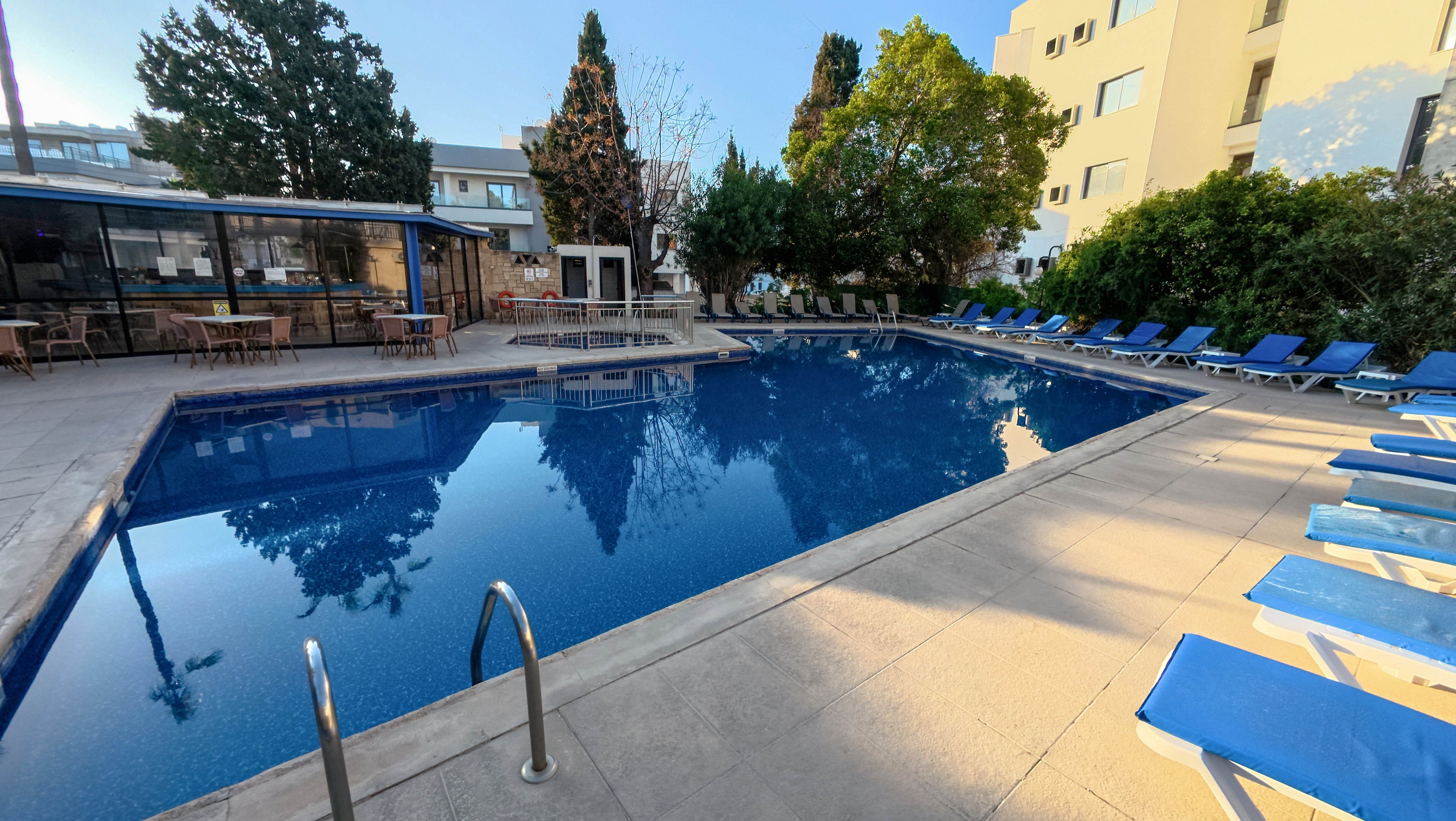 A rectangular outdoor swimming pool sits in the courtyard of a modern hotel or apartment complex. Blue lounge chairs line the poolside, suggesting a relaxing area for guests to sunbathe. Trees and shrubs surround the pool, reflecting in the calm water along with nearby buildings. The scene is brightly lit, indicating a clear day and a tranquil atmosphere.