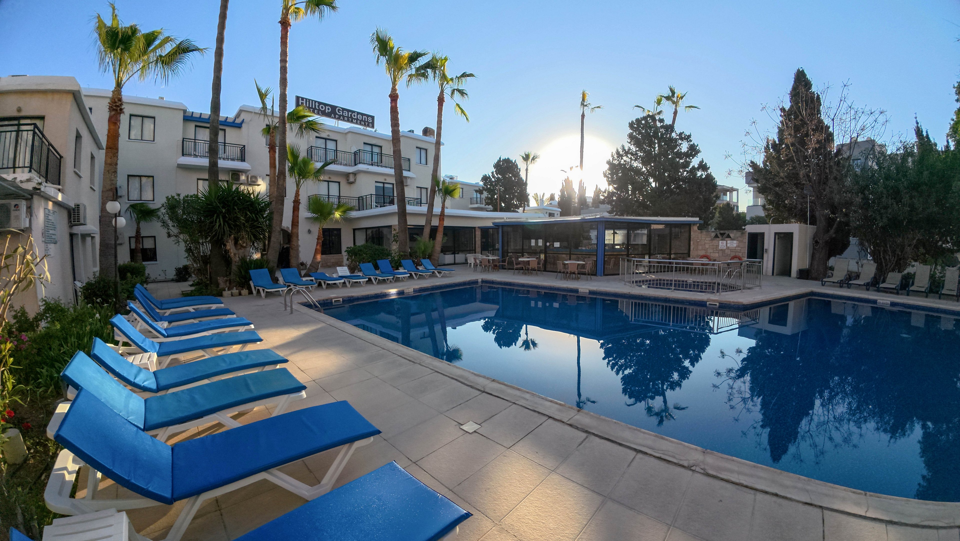A modern hotel swimming pool is surrounded by bright blue sun loungers neatly arranged along the deck. The scene is set outdoors in a courtyard framed by low-rise white buildings and tall palm trees. Calm water reflects the clear sky and surrounding architecture, creating a tranquil atmosphere. The low sun near the horizon suggests early morning or late afternoon light, enhancing the relaxed resort mood.