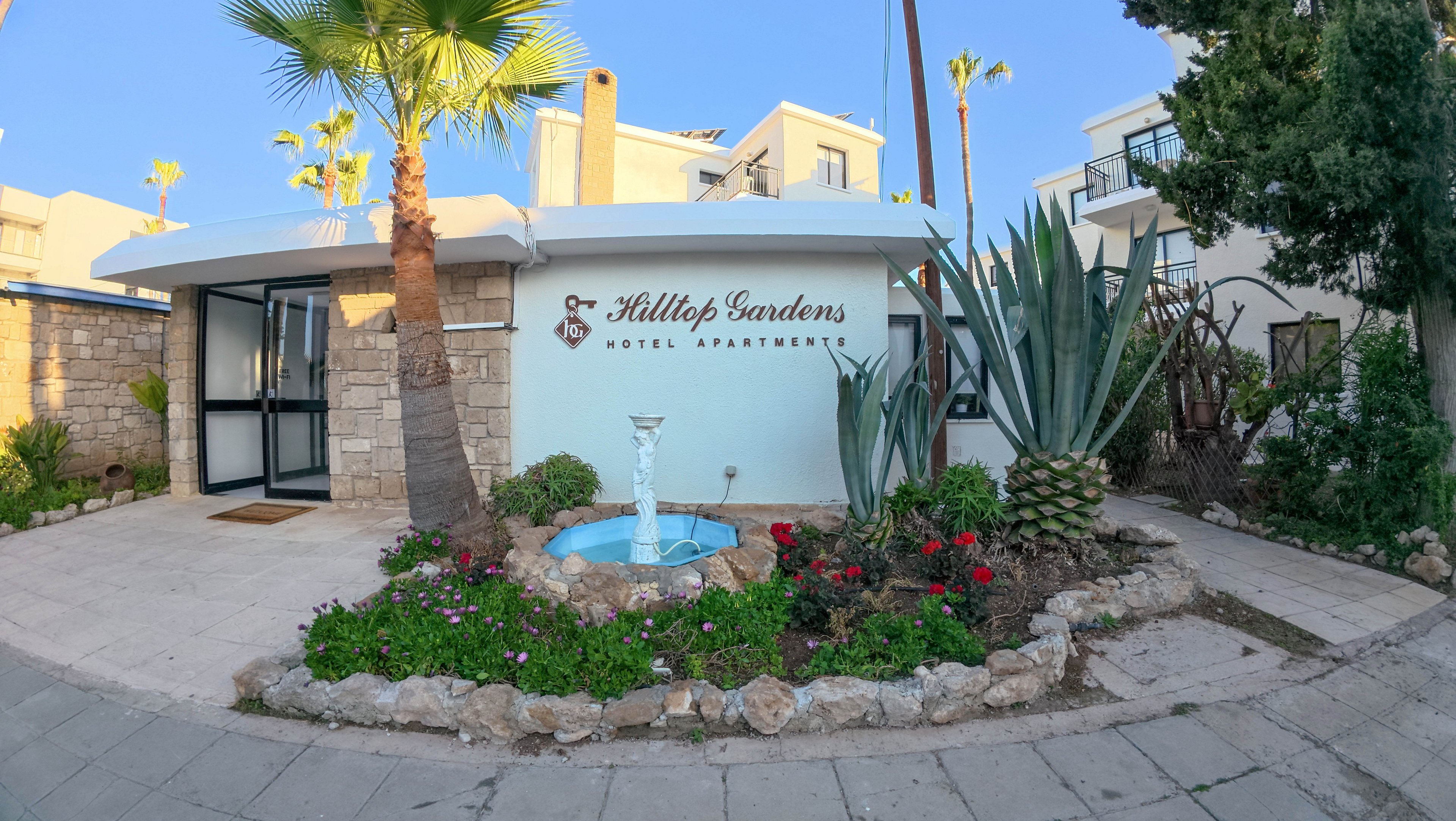 The image shows the exterior entrance of Hilltop Gardens Hotel Apartments. A low white building with stone accents faces a paved walkway, with the property name clearly displayed on the wall. In front of the entrance is a small circular fountain surrounded by a landscaped rock garden with flowers, succulents, and a large agave plant. Tall palm trees and neighboring white buildings are visible in the background under a clear blue sky, suggesting a warm, sunny climate.