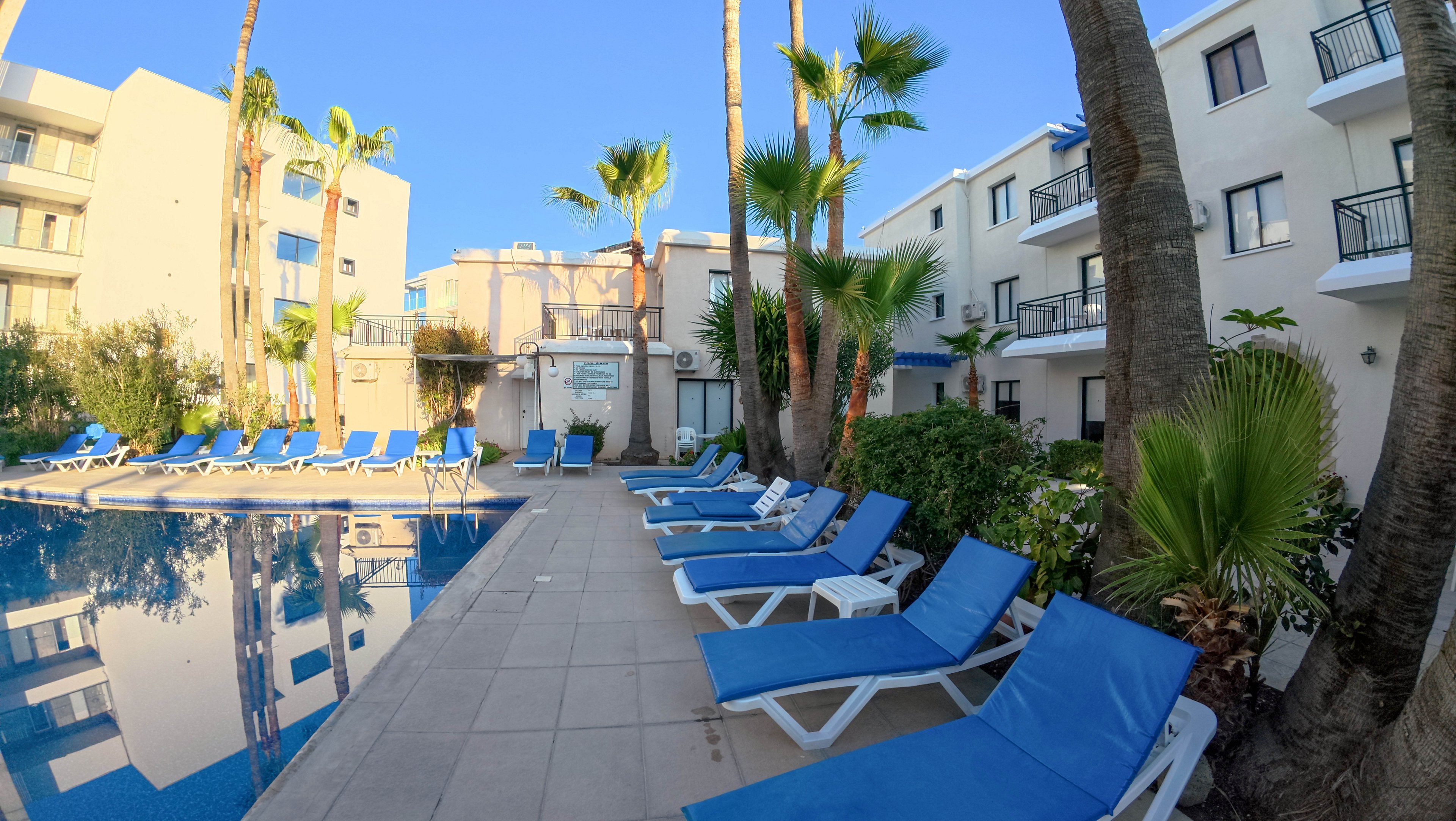 A tranquil hotel courtyard features a swimming pool lined with bright blue sun loungers. White low-rise buildings with balconies surround the pool area, creating an intimate resort atmosphere. Tall palm trees and lush greenery add a tropical feel under a clear blue sky. The scene is calm and empty, suggesting an inviting place for relaxation and sunbathing.