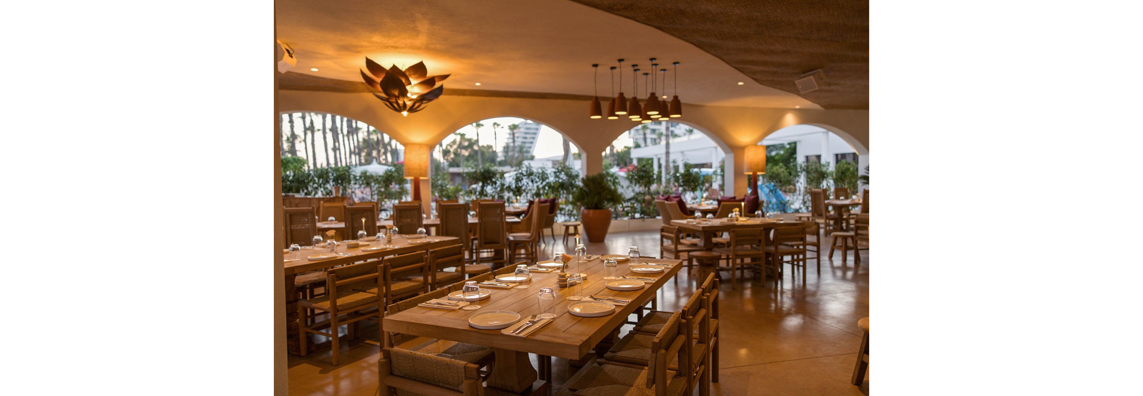 An open-air restaurant dining area is shown with wooden tables neatly set with plates, glasses, and cutlery. Soft, warm lighting from ceiling fixtures and decorative lamps creates a cozy ambiance. Large arched openings reveal greenery and an outdoor view beyond the terrace. The space appears ready for guests, emphasizing a relaxed yet elegant dining experience.