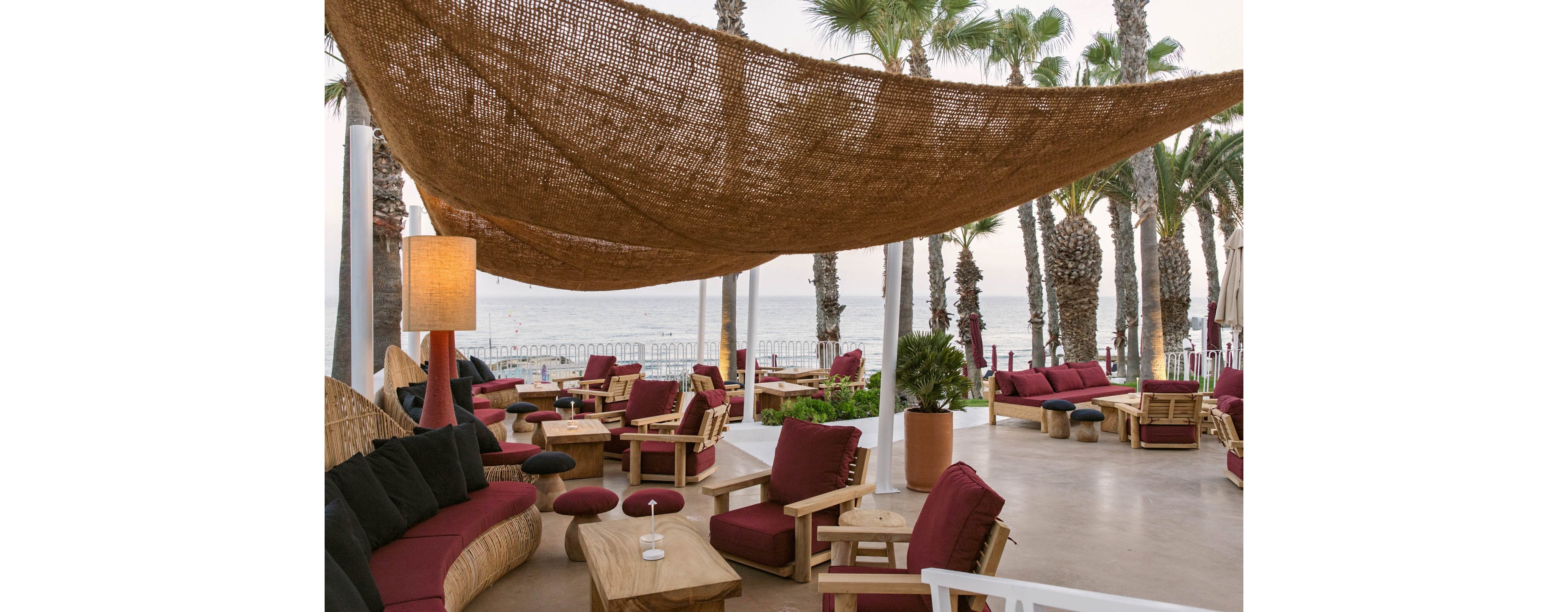 An open-air lounge area features wooden furniture with deep red cushions arranged under a large woven canopy. The seating is grouped around low tables, creating an inviting space for relaxation and conversation. Tall palm trees surround the terrace, framing a view of the ocean and shoreline in the background. Soft lighting and natural materials contribute to a warm, coastal atmosphere.