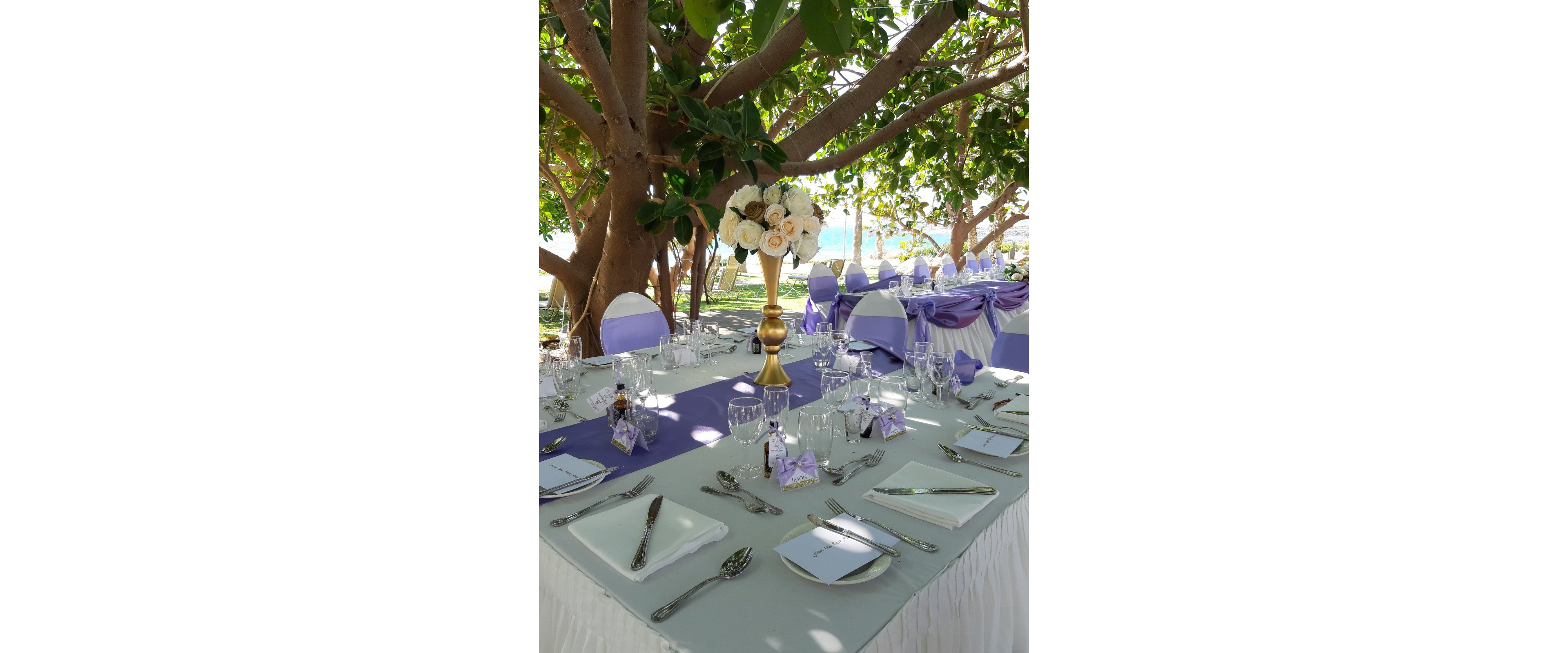 An elegantly arranged wedding reception table is set outdoors beneath lush green trees. White linens with a lavender runner and matching chair sashes create a soft, romantic color palette. A tall gold centerpiece holds a bouquet of white and cream roses, surrounded by neatly arranged glassware, plates, and cutlery. Place cards and small favor boxes add personalized details to the formal table setting.