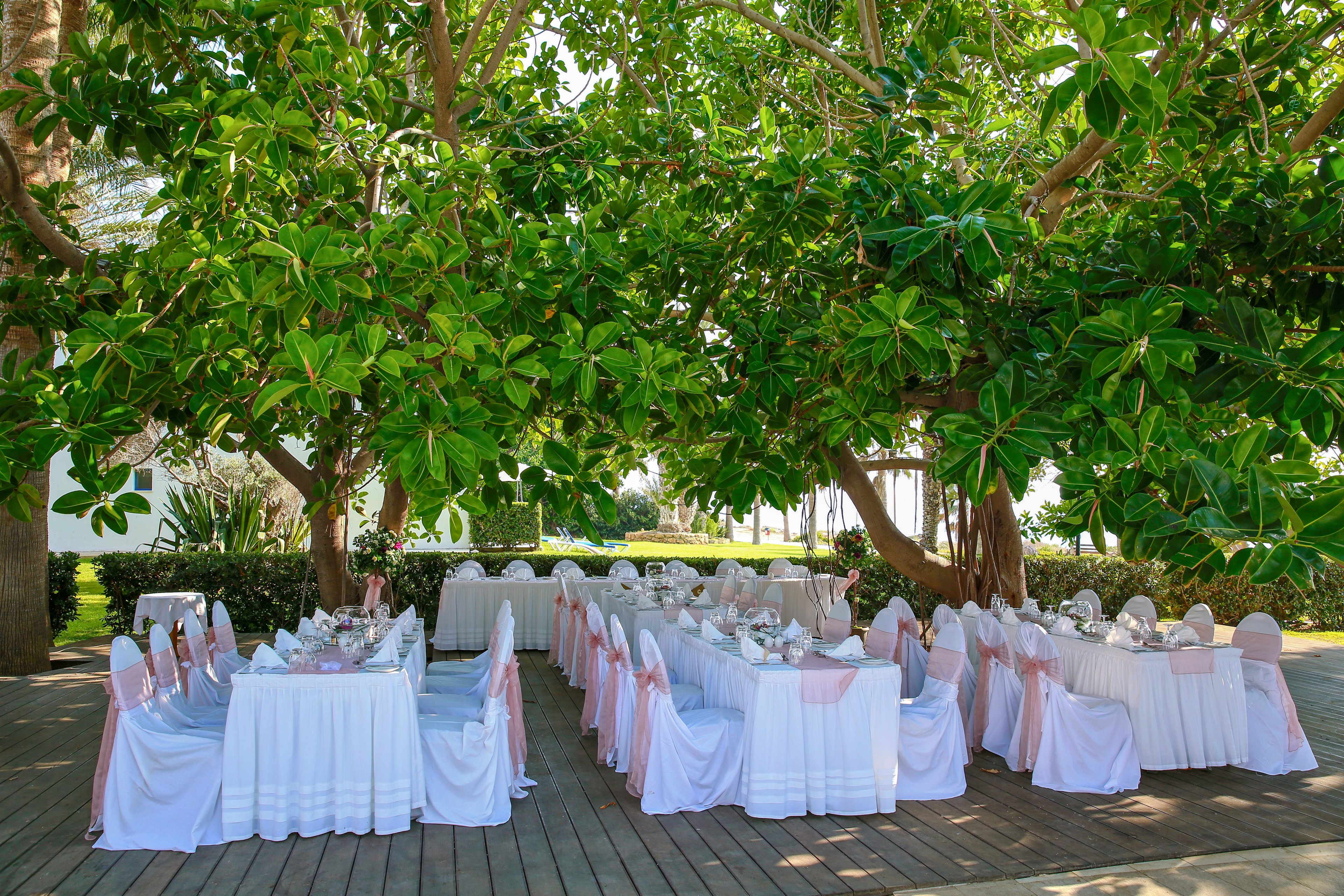 An elegant outdoor wedding reception is arranged beneath large, leafy trees providing natural shade. Long rectangular tables are covered with white linens and set with glassware, plates, and folded napkins. White chairs with soft pink sashes line both sides of the tables, creating a romantic and formal atmosphere. The wooden deck flooring and surrounding greenery suggest a garden or resort setting prepared for guests.