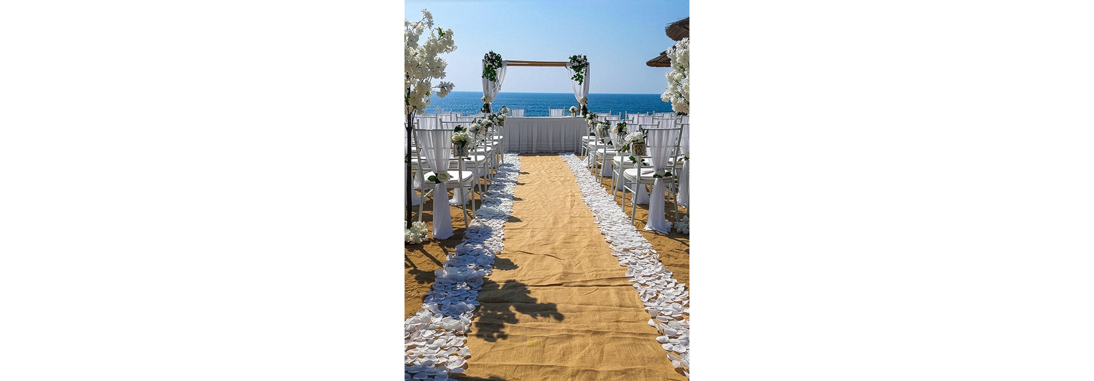 A wedding ceremony setup is arranged on a sandy beach facing the ocean. A burlap-style aisle runner is lined with white flower petals leading to a simple wooden arch decorated with white drapery and greenery. White chairs with floral arrangements flank the aisle on both sides, creating a symmetrical composition. The bright blue sea and clear sky provide a serene, romantic backdrop for the outdoor event.