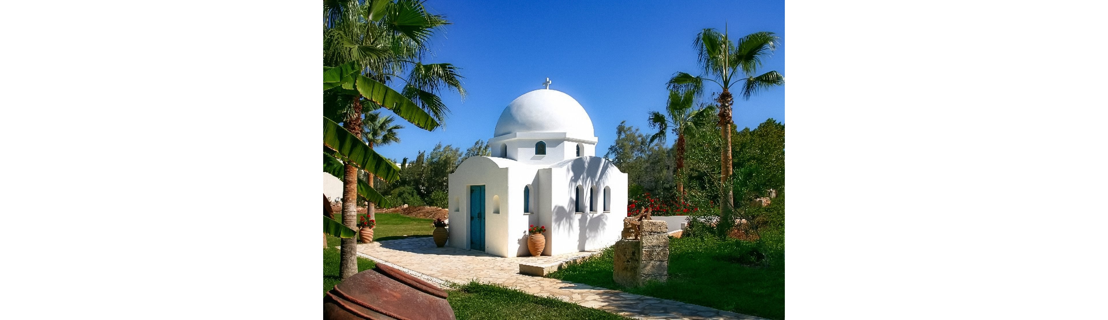 A small white chapel with a rounded dome stands in a lush garden setting. The building features arched windows, a light blue door, and simple architectural lines typical of Mediterranean or Greek island style. Palm trees and terracotta pots surround the chapel, adding to the tranquil, sunlit atmosphere. A bright blue sky and vibrant greenery emphasize the serene, vacation-like mood of the scene.