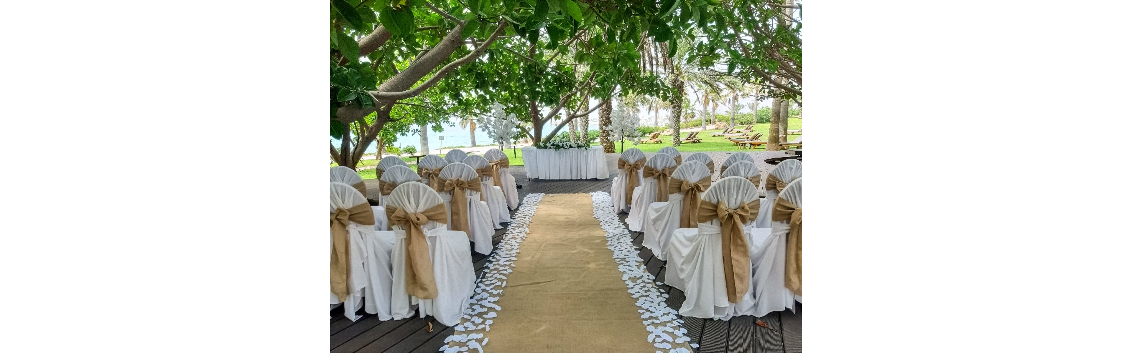 An outdoor wedding ceremony setup features rows of white-covered chairs tied with rustic burlap bows. A beige aisle runner stretches down the center, bordered by scattered white flower petals. The scene is shaded by lush green trees, with glimpses of palm trees and lawn chairs in the background. A decorated table or altar stands at the end of the aisle, ready for the ceremony.