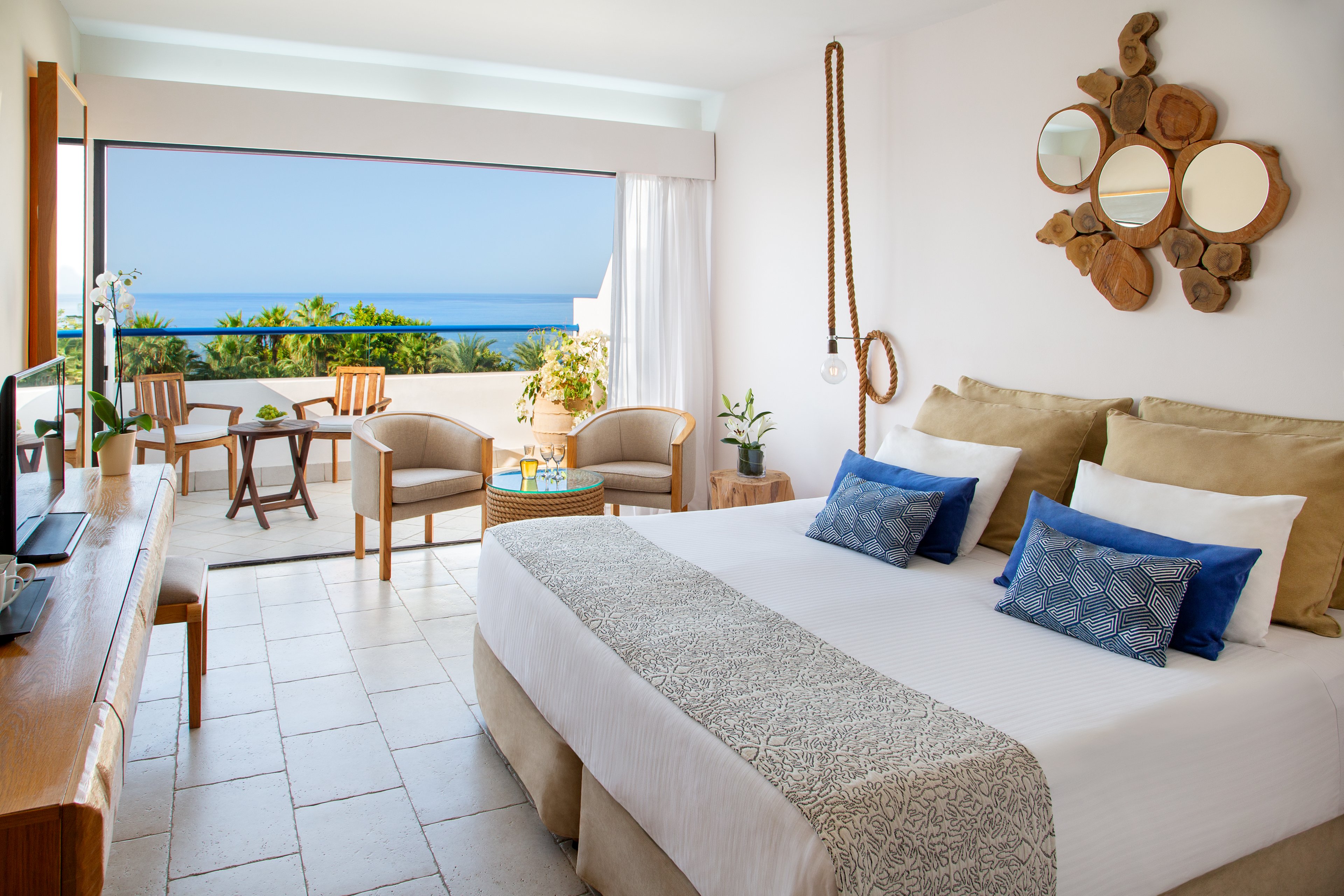 A modern hotel bedroom features a large neatly made bed with neutral linens and blue accent pillows. The room opens onto a balcony with chairs and a small table overlooking a clear blue sea and distant horizon. Natural light floods the white interior, highlighting wooden furniture and decorative mirrors on the wall. The overall atmosphere is calm, airy, and inviting, ideal for a relaxing seaside vacation.