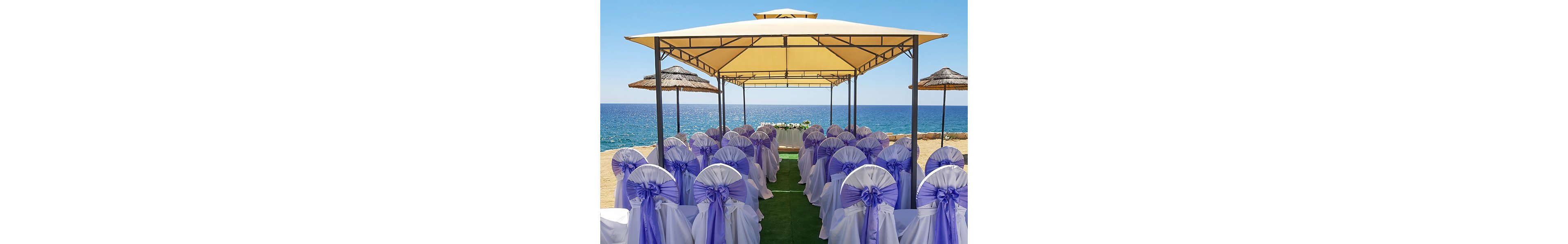 Rows of white chairs with lavender bows are arranged under a canopy for an outdoor wedding ceremony. The setup faces a small altar decorated with flowers at the end of the aisle. Beyond the structure, the blue sea and clear sky create a bright coastal backdrop. Straw parasols and sandy ground emphasize the beachside location and relaxed yet elegant atmosphere.