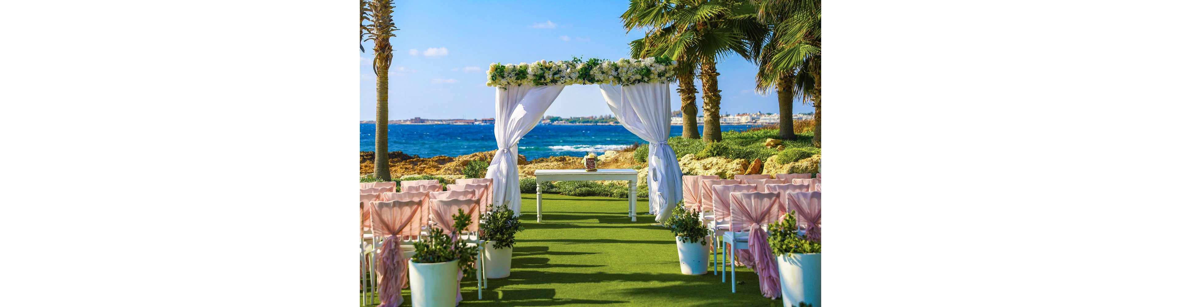 An outdoor wedding ceremony is arranged on a lush green lawn overlooking a bright blue sea. Rows of chairs with soft pink fabric bows face a white archway draped in flowing curtains and topped with fresh flowers. A simple white table stands beneath the floral arch, framed by potted greenery and palm trees. The clear sky and vibrant colors create a cheerful, romantic atmosphere by the water.