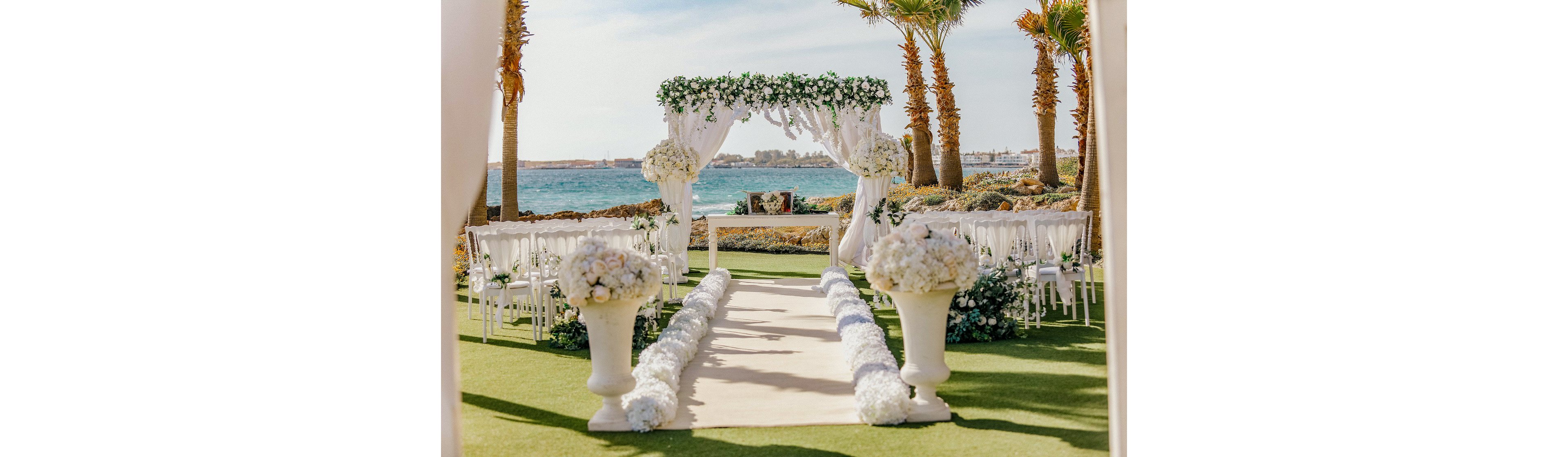 An elegant wedding ceremony setup is arranged outdoors beside a bright blue sea. A white fabric-draped arch decorated with lush green foliage and white flowers frames the altar area. Rows of white chairs line a central aisle bordered by large floral arrangements and spherical bouquets. Palm trees and a sunny sky create a tropical, romantic atmosphere.