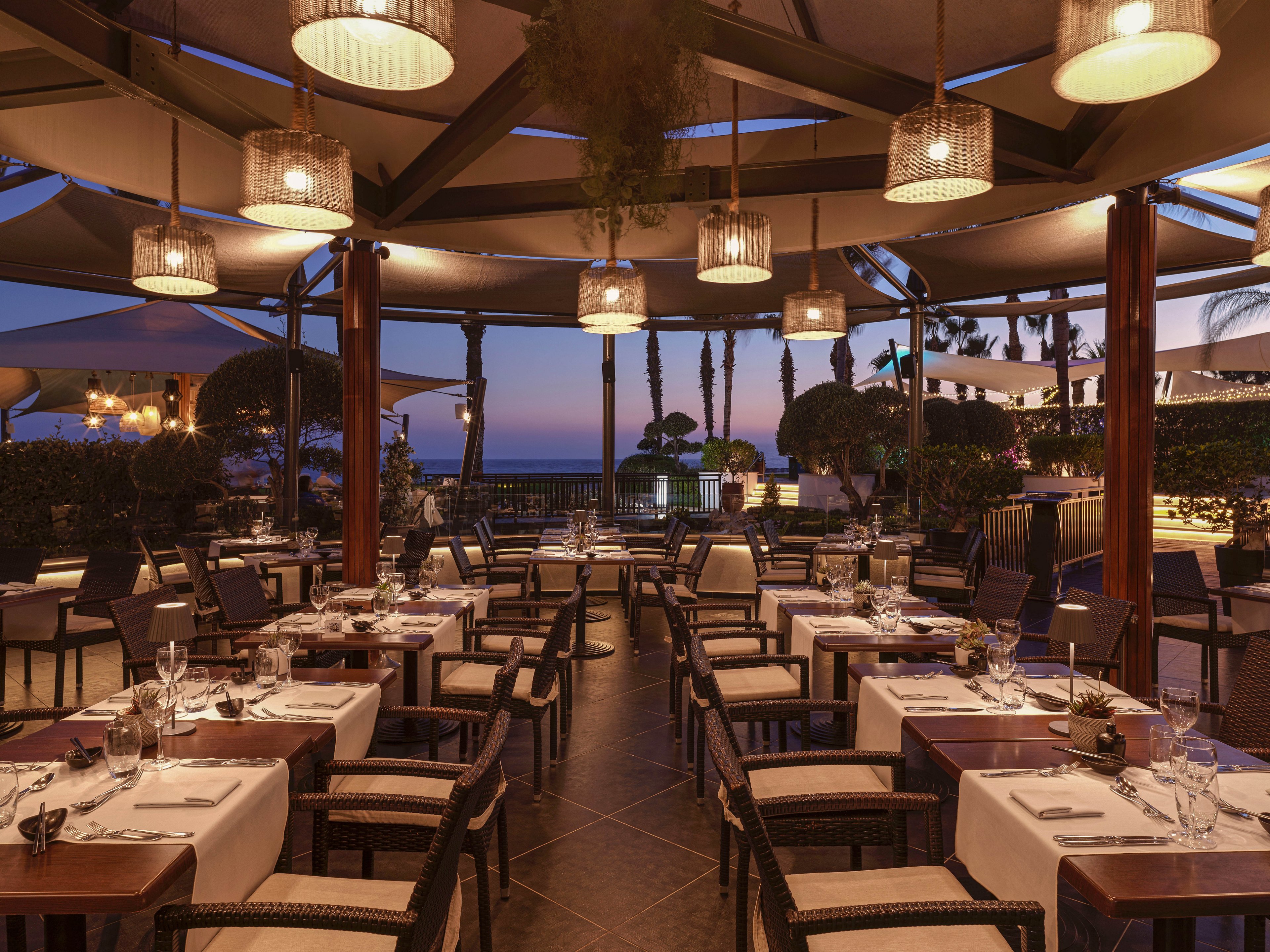 An upscale open-air restaurant is set for evening dining with neatly arranged tables and white tablecloths. The space features a covered terrace structure with hanging pendant lights casting a warm glow. Beyond the seating area, lush greenery and palm trees frame a distant view of the sea at twilight. The overall atmosphere is calm, sophisticated, and inviting for guests.